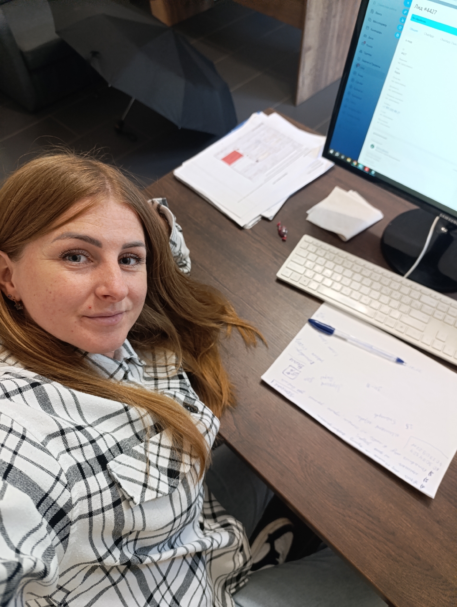 A woman with long, reddish-brown hair, wearing a black and white plaid shirt, is lying down in an office setting, looking at the camera. She has fair skin, freckles, and light-colored eyes. Her hair is spread out around her. In the background, a computer monitor displays a software interface, a white keyboard, a stack of papers, and a folded black umbrella. The scene appears to be a desk area with wooden flooring.