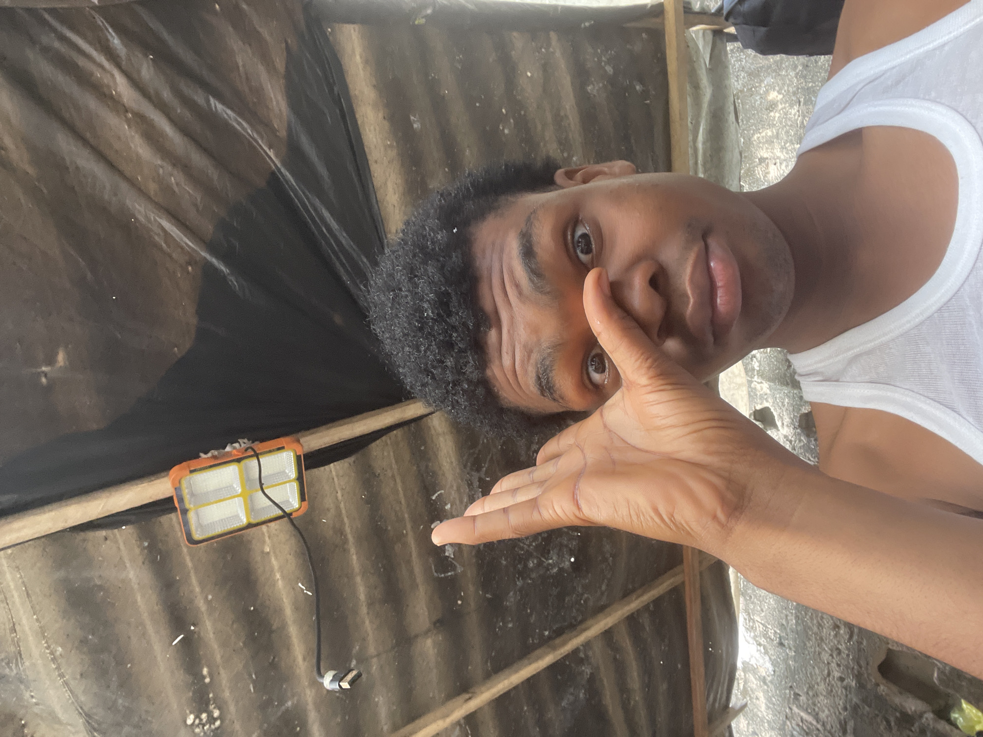 In Ilado, Nigeria, a young Black male wearing a white tank top poses for a selfie under a corrugated metal roof supported by wooden beams. A black tarp is stretched behind him, and an orange and yellow work light hangs from the ceiling. His right hand is raised, obscuring his left eye and nose, while his right eye is visible and wide. The background appears to be an outdoor, possibly under-construction or storage area.
