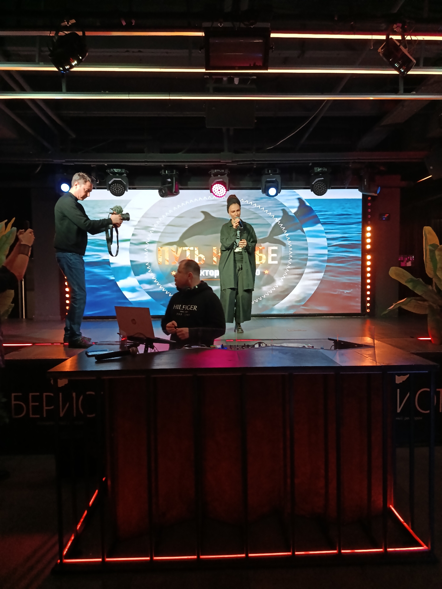 The image depicts an event taking place in a dim, indoor venue. A woman stands on a stage, holding a microphone and appearing to speak. Behind her, a large screen displays an image of dolphins swimming in the ocean, with some text overlaid. To the left of the stage, a man stands, holding a camera and aiming it at the woman. In the foreground, a desk with a laptop sits, with another man seated behind it. The environment is dark, with various lighting fixtures illuminating the scene. Some signage is visible in the foreground, though the details are obscured. The location is identified as the city of Moscow, Russia.