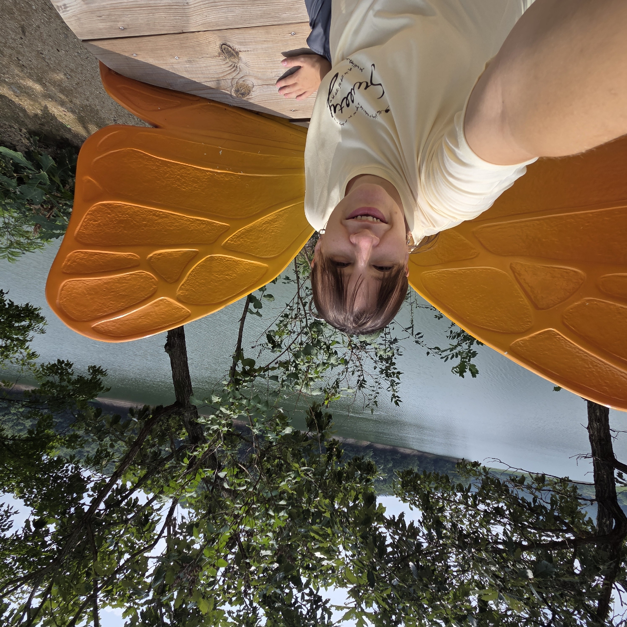 A woman with brown hair and bangs is taking a selfie outdoors. She is smiling and wearing a light yellow t-shirt with a heart-shaped graphic that says "Sweetty" in cursive. She has large, orange, butterfly-like wings attached to her back. She is sitting on a wooden deck next to a body of water, likely a lake or reservoir. The background consists of lush green trees and a cloudy sky. The location is Gapcheon-myeon, South Korea.