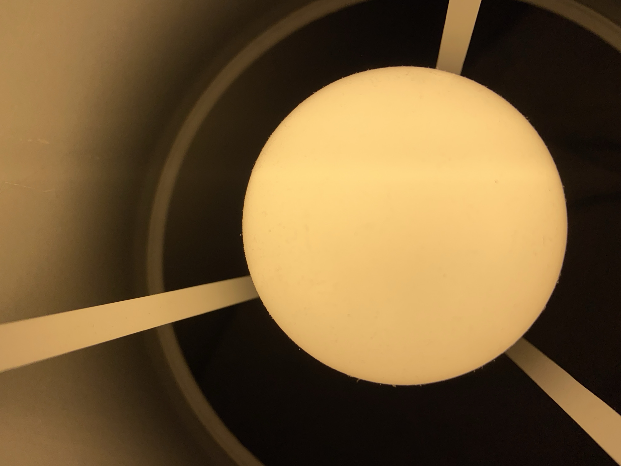 An overhead view captures the interior of a light fixture, situated in Budva, Montenegro. The central and dominant subject is a spherical, off-white to pale yellow luminous object, consistent with an illuminated light bulb or diffuser. Its surface exhibits minor textural irregularities. Three narrow, planar strips of a similar off-white or beige hue are visible, appearing to function as structural supports or framing elements for the spherical light. Two strips extend from the upper periphery of the frame towards the sphere, while a third is present in the lower-left. The immediate background is a dark, curved surface, suggesting the interior of a cylindrical or conical lampshade, which encloses the light source and its supports. No individuals, direct actions, or interactions are visible.