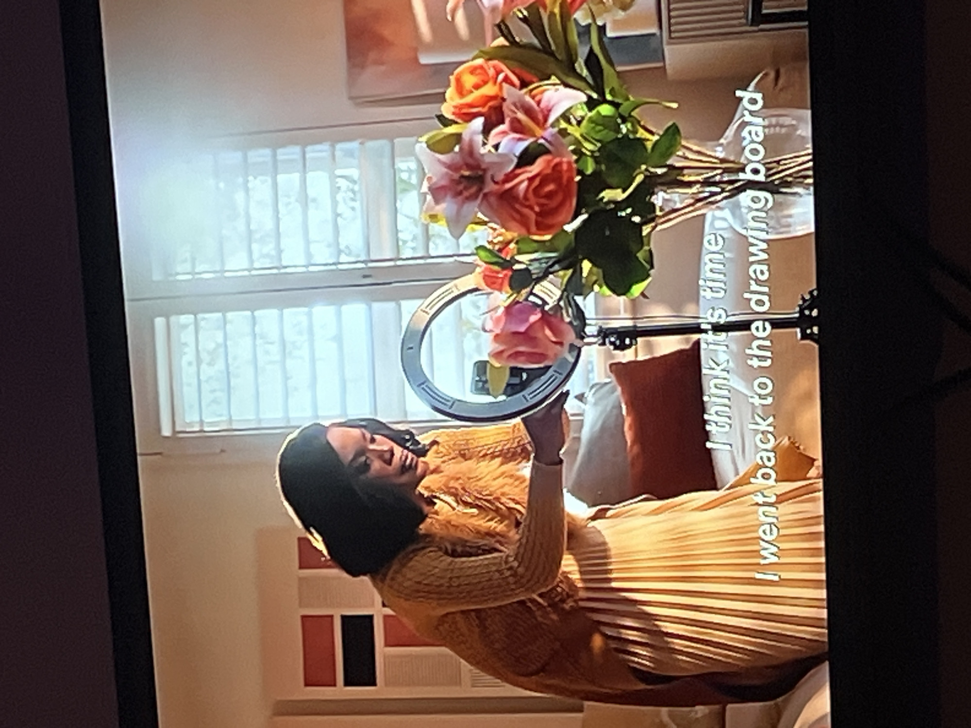 This media file depicts a person in an indoor setting. The subject appears to be a woman with dark hair, wearing a yellow textured sweater and a pleated skirt. She is holding a ring light, suggesting she might be recording content or taking photos.
Behind the woman, there is a bouquet of flowers with various colors, including pink, orange, and white. The background includes a window with blinds, indicating it might be daytime. There is also a glimpse of furniture, including pillows and what appears to be a vase. The overall scene suggests a creative or artistic environment.
Visible text overlaid on the image reads: "I think it's time I went back to the drawing board". This text appears to be a caption or a quote related to the visual content.
The image quality is not high, and the orientation is rotated, making precise details difficult to discern. No specific information about the time of day, weather, or emotions beyond a neutral or contemplative expression on the subject's face can be determined. The provided location context of Gura Top, Nigeria, cannot be visually confirmed from the image itself.