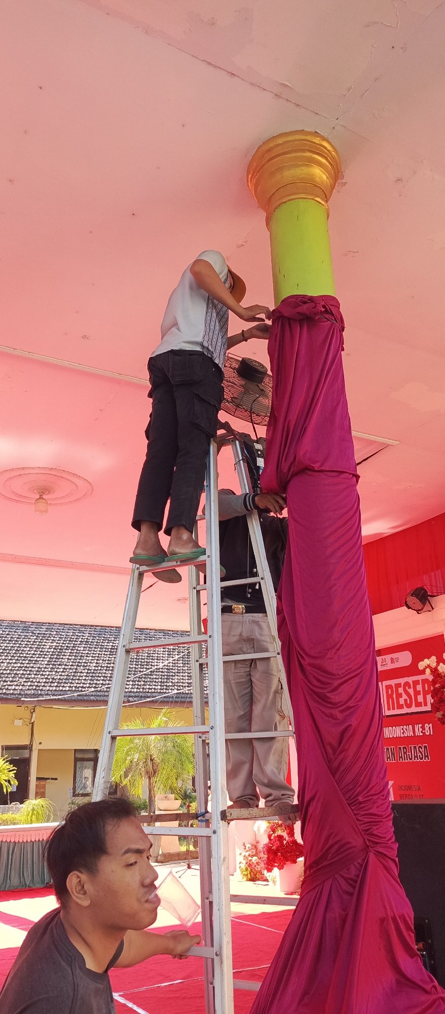 In Arjasa, Indonesia, two individuals are actively engaged in setting up decorations around a large, green pillar adorned with a golden cap. One person, positioned higher on a silver ladder, wears a white and blue striped shirt, a light-colored cap, and dark pants. They are observed adjusting the rich, magenta fabric that drapes elegantly down the pillar, seemingly securing it. A fan is attached to the ladder, possibly for ventilation. Below them, another person, clad in a grey jacket and light-colored pants, stands on the ladder, also appearing to be involved in the decorative arrangement. In the foreground, a man with short dark hair is captured in profile, holding a clear plastic cup and looking towards the pillar. The setting suggests an outdoor event or preparation for one, with a pink ceiling overhead, architectural elements visible in the background, and a red carpet laid out on the ground.