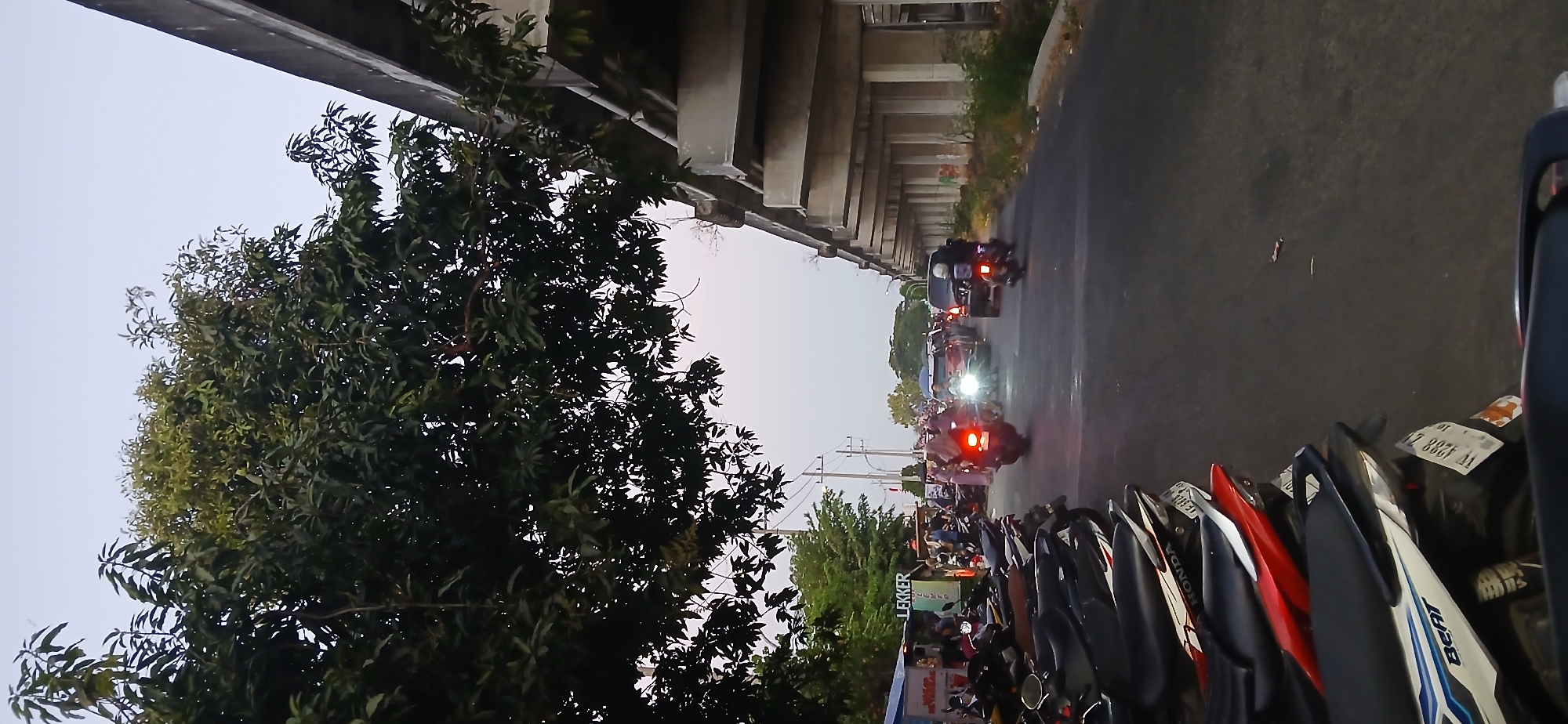 In Wadungasri, Indonesia, a street scene unfolds under an overpass. The foreground is dominated by a row of parked motorcycles, their seats a mix of black, red, and white. Beyond them, the asphalt road shows signs of traffic. A motorcycle with its brake light illuminated travels away from the viewer, followed by a car with its headlights on. Further down the road, more vehicles are visible, including a black SUV and another motorcycle. On the left side of the street, a small market stall with a "LEKKER" sign is situated, with several people gathered around it. The overpass, constructed of concrete pillars and beams, casts a shadow over the right side of the scene. Lush green trees line the left side of the road, their branches reaching towards the overcast sky. The overall atmosphere suggests a busy, everyday street in a Southeast Asian urban or suburban setting.