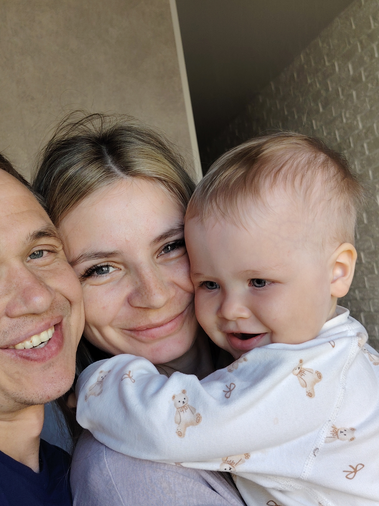 The image shows three people posing closely together for a selfie, potentially in a residential setting. On the left, a man with fair skin and short hair is smiling broadly, revealing his teeth. To his right, a woman with fair skin and freckles is also smiling, with her head positioned near the man. She appears to be holding a baby with fair skin and light-colored hair. The baby is turned towards the right side of the frame, also smiling. The baby is wearing a white long-sleeved garment decorated with teddy bear and bow tie patterns. The background appears to be a textured wall, and a portion of a window or doorway is visible on the left. There is no evidence to confirm or deny that the image was taken in the city of Chaikovsky, Russia.