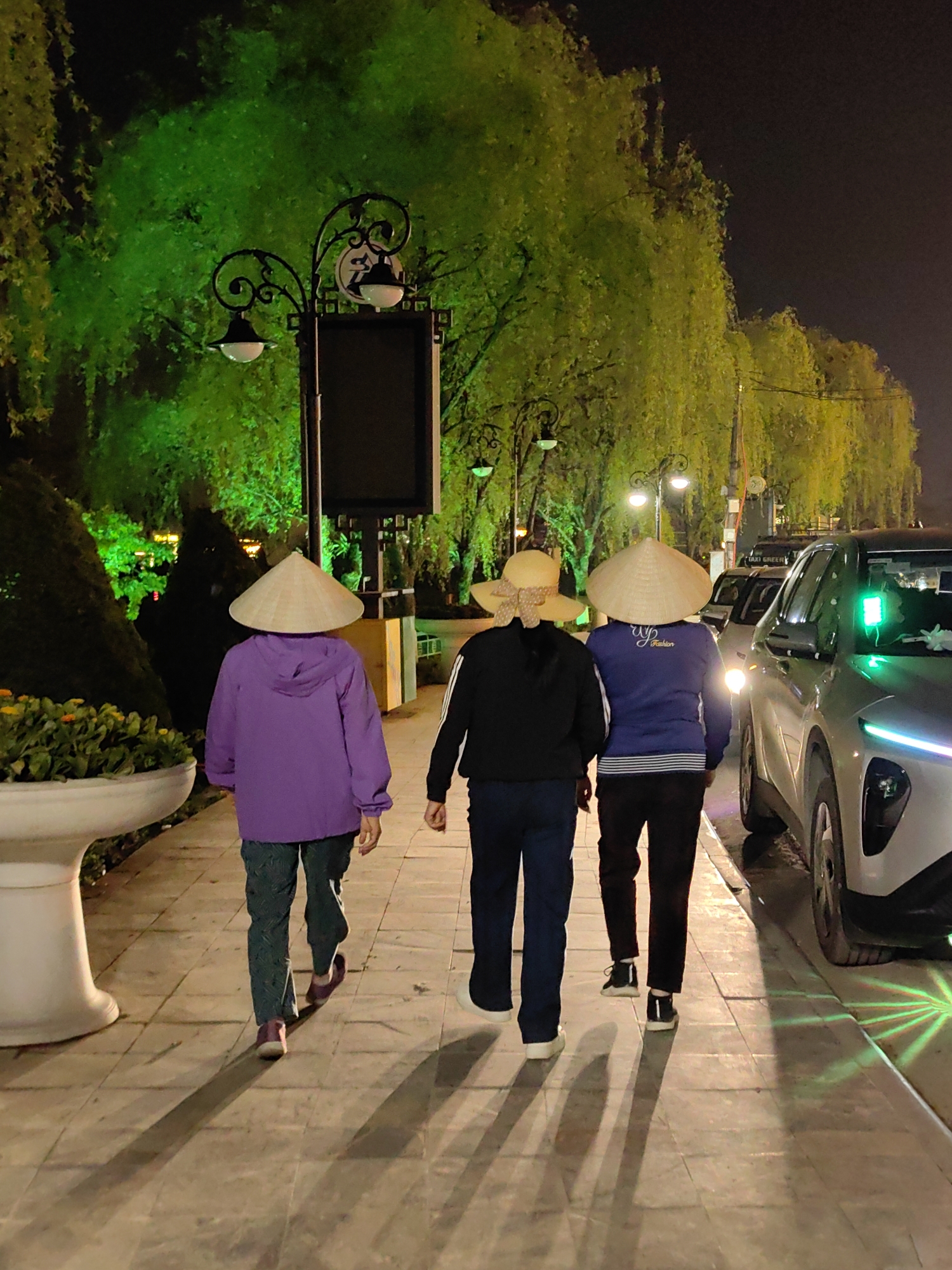 At night in Sa Pa, Vietnam, three individuals walk away from the camera on a paved sidewalk. The person on the left wears a purple hooded jacket, dark patterned pants, purple shoes, and a traditional conical nón lá hat. The middle person wears a black jacket with white arm stripes, dark pants, white sneakers, and a light-colored straw hat with a patterned bow. The person on the right wears a dark blue long-sleeved shirt with "WY Fashion" text, dark pants, black sneakers, and a traditional conical nón lá hat. Their long shadows are cast on the light-colored sidewalk.
To their right, a light-colored vehicle with illuminated headlights and a blue LED strip is parked or moving slowly on the adjacent road. Another vehicle and a "TAXI GREEN" sign are visible further down the road. The street is lined with ornate black lampposts featuring globe lights; one includes a dark rectangular sign. Lush trees in the background are dramatically illuminated with green light. On the left, a white, ornate planter contains green foliage and yellow flowers, next to a trimmed conical hedge. The sky above is dark.