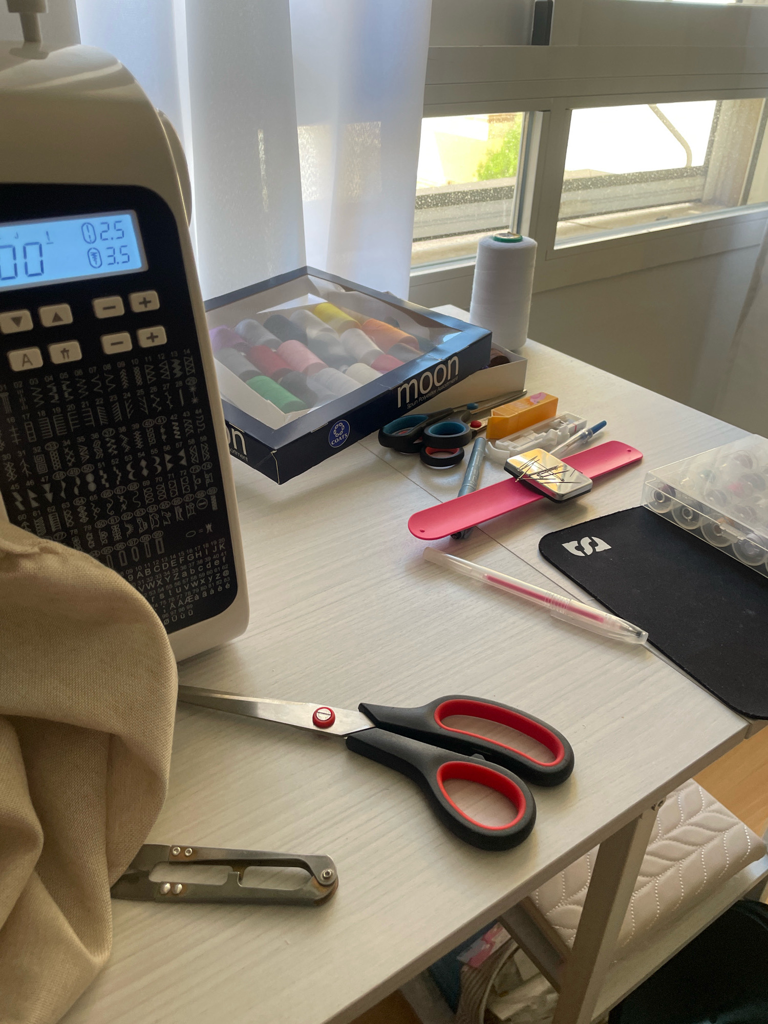 The image depicts a sewing machine and various sewing supplies arranged on a white wooden table. The sewing machine, with a digital display showing "00" and settings "2.5" and "3.5", is positioned on the left side of the frame. To the right of the sewing machine, a box of colorful sewing threads is visible, along with a spool of white thread. Other sewing accessories, including scissors, a pink magnetic wrist pin holder with pins, a seam ripper, a clear box of bobbins, and a pink pen, are also present on the table. A beige fabric is draped over part of the sewing machine. In the background, a window with white curtains allows natural light to illuminate the scene. The overall environment suggests a home sewing area. The location is identified as Atarfe, Spain.