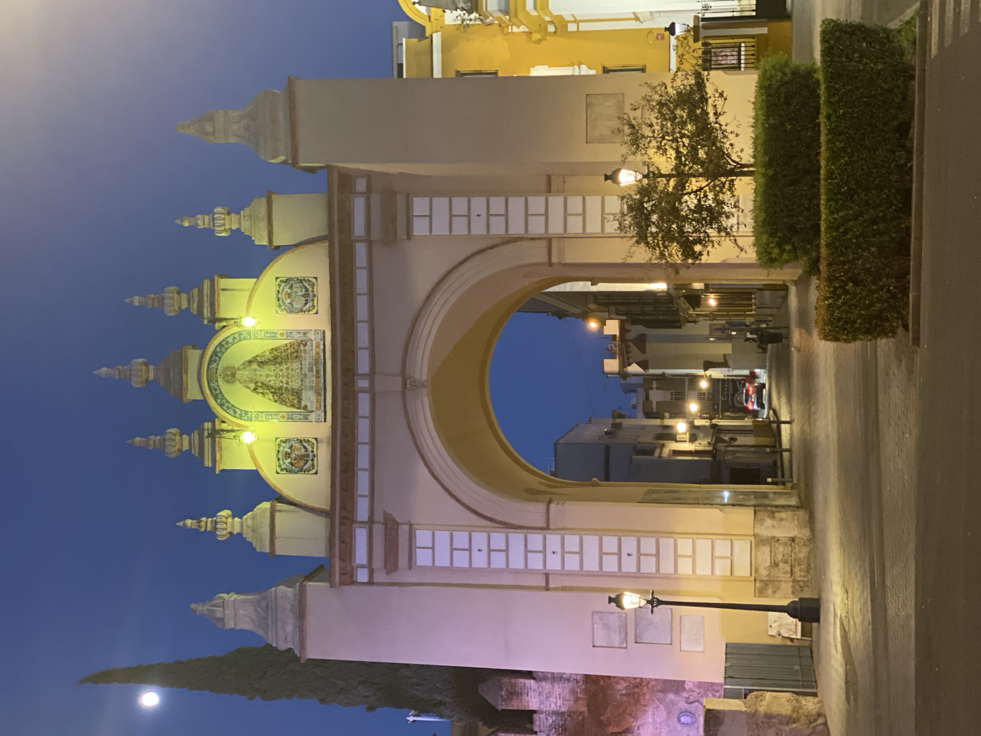 The image shows the Puerta de Jerez, a monumental archway in Seville, Spain, at twilight. The arch is illuminated, highlighting its ornate details, including ceramic tiles depicting a religious figure at the top. The moon is visible in the dark blue sky to the left of the arch, next to a tall, slender tree. Streetlights are on, casting pools of light on the cobblestone street and illuminating the facade of the arch.
Through the archway, a narrow street recedes into the distance, lined with buildings in shades of yellow and cream, also lit by artificial lights. A red car is visible driving away from the viewer down the street. One person is partially visible walking on the sidewalk to the right of the arch. In the foreground, to the right of the arch, are two neatly trimmed hedges. Another lamppost is visible on the right side of the image, and a portion of a yellow building with decorative ironwork is visible on the far right. The overall scene is one of a well-lit urban environment at dusk, emphasizing the architectural beauty of the Puerta de Jerez and its surroundings.