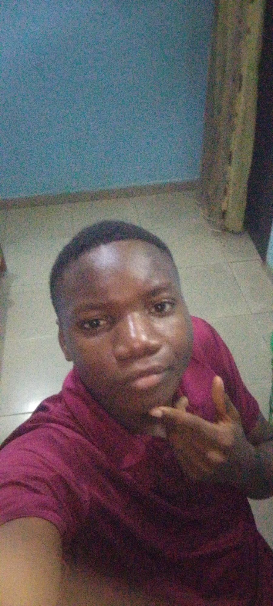 A selfie is taken from a low angle of a young Black male. He is wearing a maroon collared shirt. His right hand is raised to his chin, with his index finger and thumb extended, and his other fingers are curled. He has short black hair and dark eyes. The background appears to be an indoor setting with a light blue wall behind him and a tiled floor beneath him. To the right of the image, a vertical wooden panel is visible.