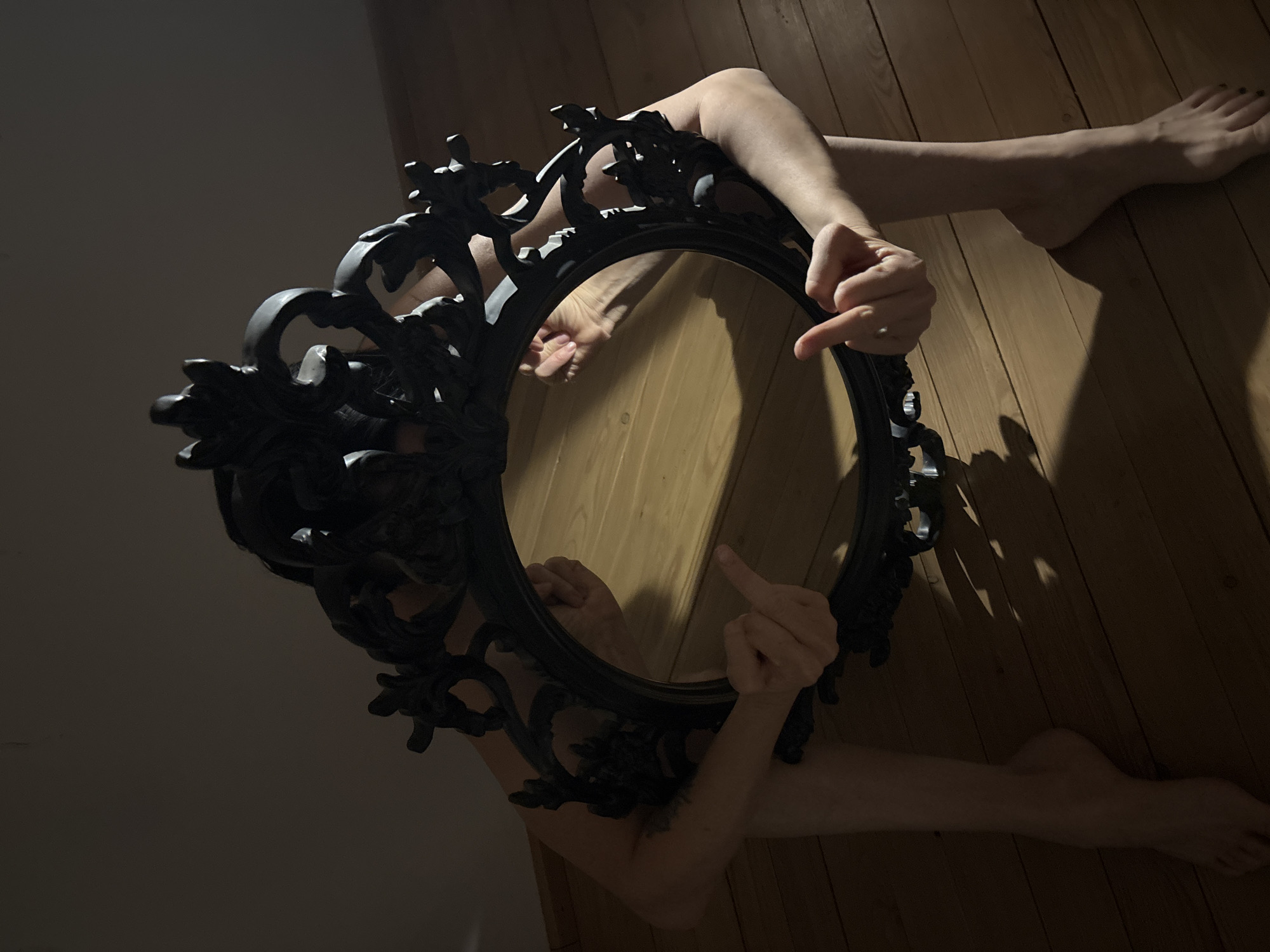 A nude person is lying on a wooden floor, partially obscured by an ornate, black-framed circular mirror. The person is holding the mirror up with their arms, revealing their reflection. One hand is giving the middle finger to the mirror. The location cannot be determined as Chaikovsky, Russia.