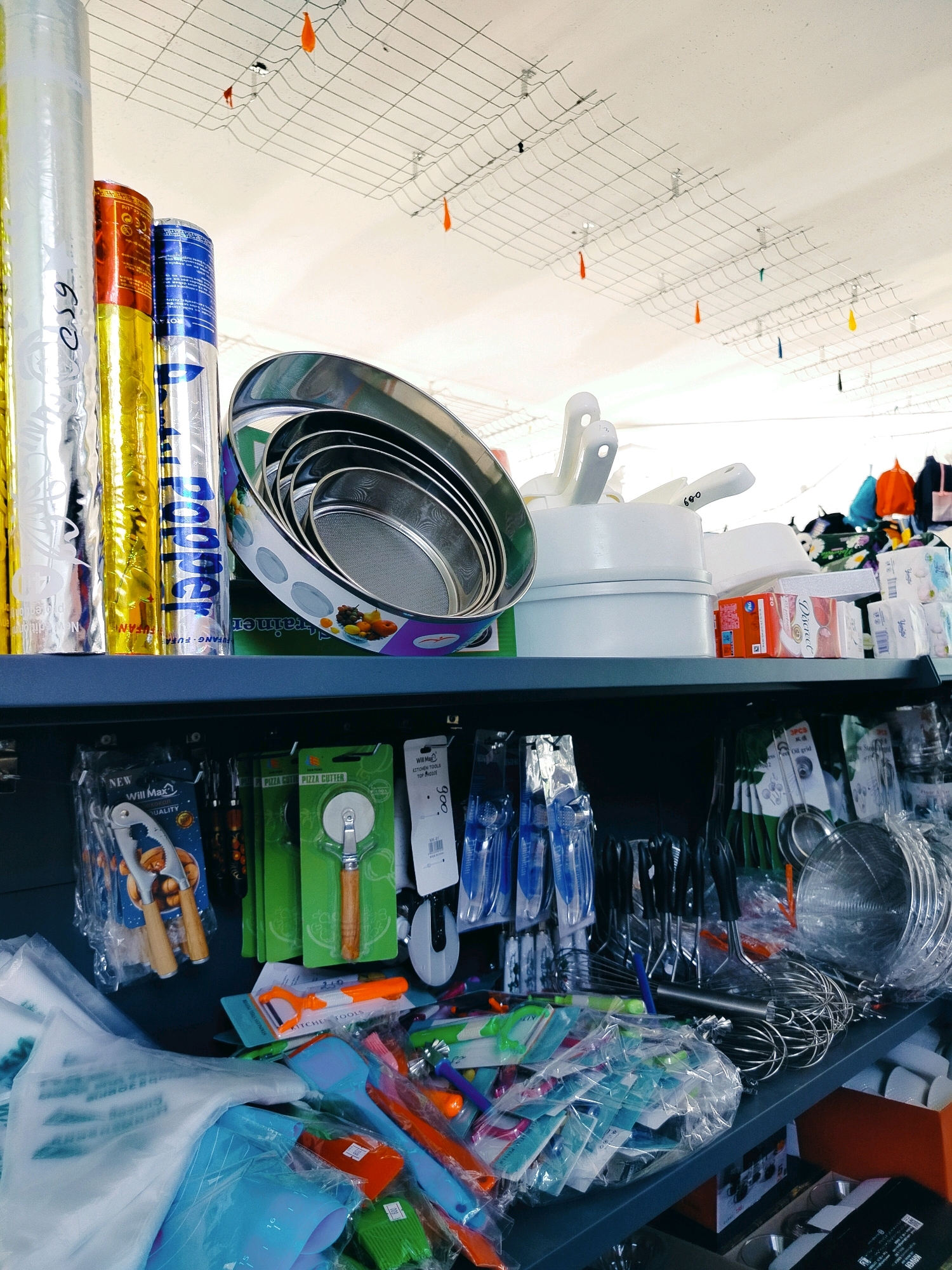 The image shows a shelf display of kitchenware in a store. On the top shelf, there are several rolls of foil or plastic wrap, a set of metal strainers stacked inside each other, and some white plastic containers and lids. In the background, colorful clothing items are visible. The ceiling has a wire grid with small orange and yellow tags hanging from it.
The second shelf is packed with various kitchen utensils and tools. On the left, there is a nutcracker in its packaging. Next to it are green-packaged pizza cutters. Further to the right are various smaller utensils like peelers and brushes, many of them bundled in plastic. A large metal whisk is also visible, along with other metal cooking tools. In the lower right corner, a cardboard box is partially visible.
The overall impression is of a retail environment, likely a kitchenware or household goods store. The lighting is bright, suggesting an indoor setting. The city of Көлкент, Kazakhstan is mentioned as the location, implying this is a local store within that city. There are no people visible in the image.
