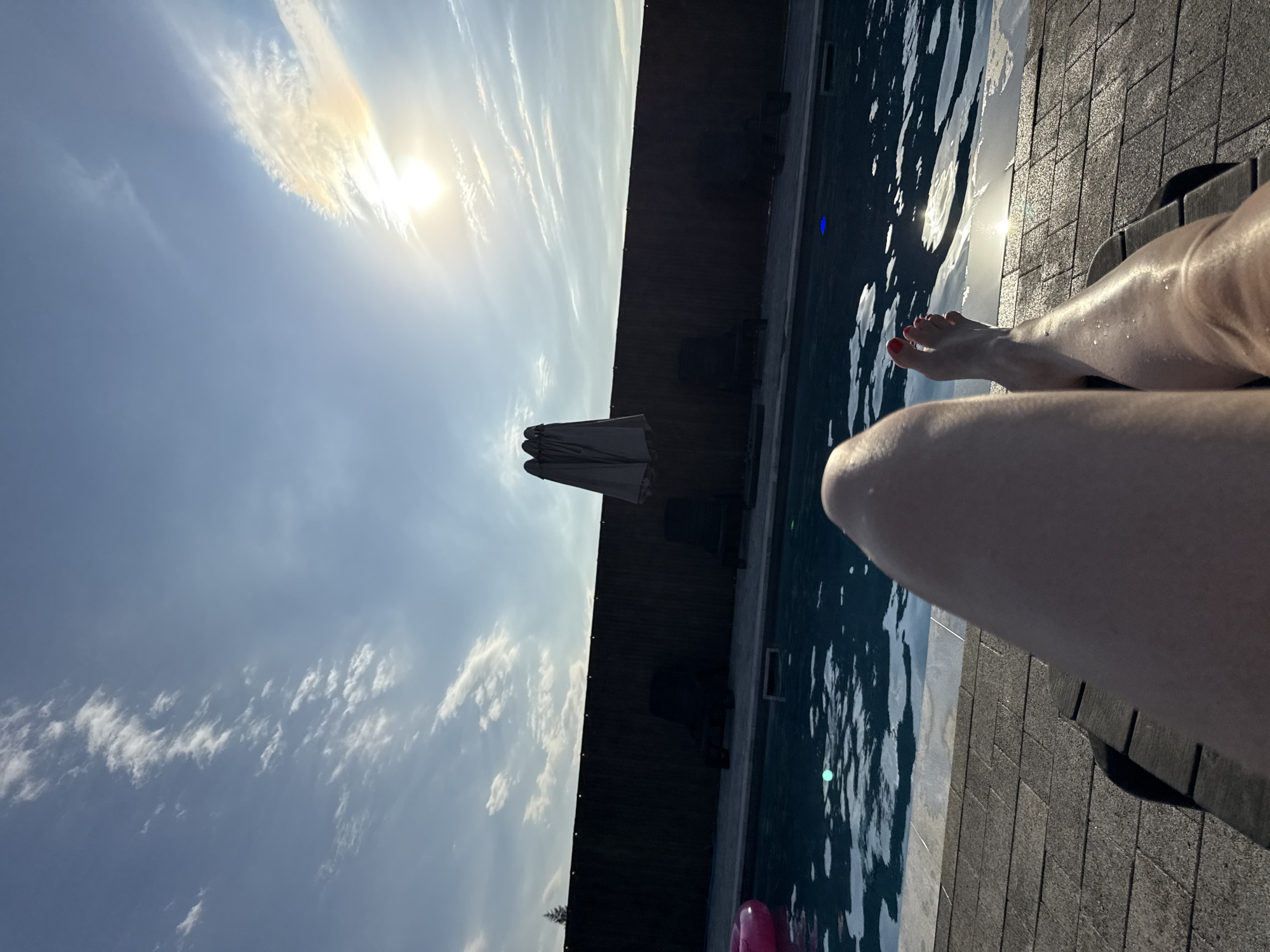 A first-person perspective shot shows a person's legs and feet, with red painted toenails, resting on a dark wooden lounger. The lounger is positioned on a textured, gray tiled surface. To the left, a swimming pool with dark blue water reflects the bright sky. The water has white ripples and a few green and blue lights. A partially visible pink inflatable ring is in the pool. A dark wooden fence runs along the back of the pool area, with three closed, light gray umbrellas standing near the fence. The sun is visible in the upper right corner, partially obscured by wispy white clouds. The sky is a clear blue. The setting appears to be outdoors, likely a backyard or resort, and is not specifically identifiable as Pervorechenskoye, Russia.