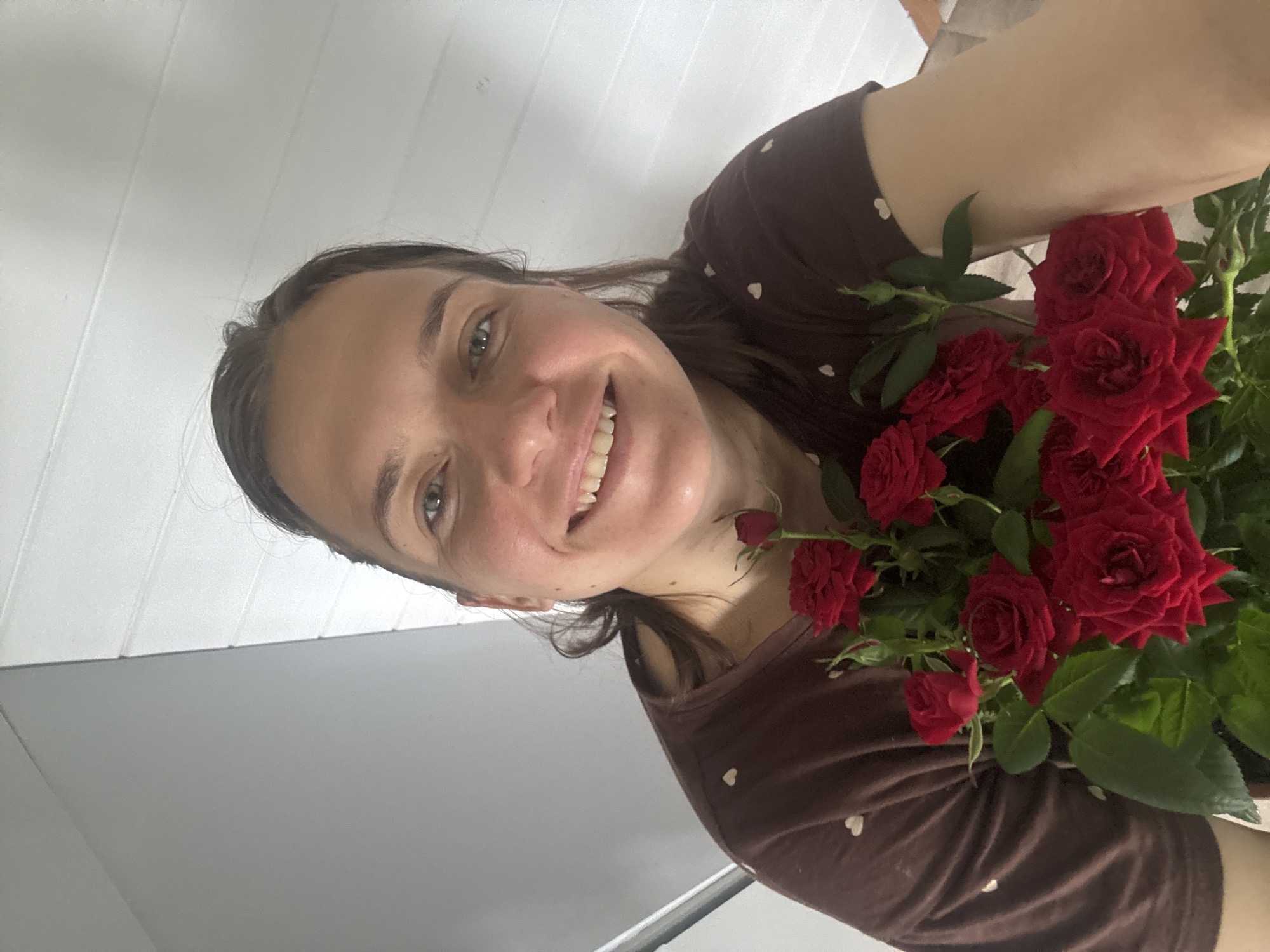 A woman is holding a bouquet of red roses. She is indoors, possibly in a room with a white ceiling. The woman has fair skin, brown hair, and is smiling. She is wearing a brown shirt with a small white heart pattern. The location is specified as Раздолье, Russia.