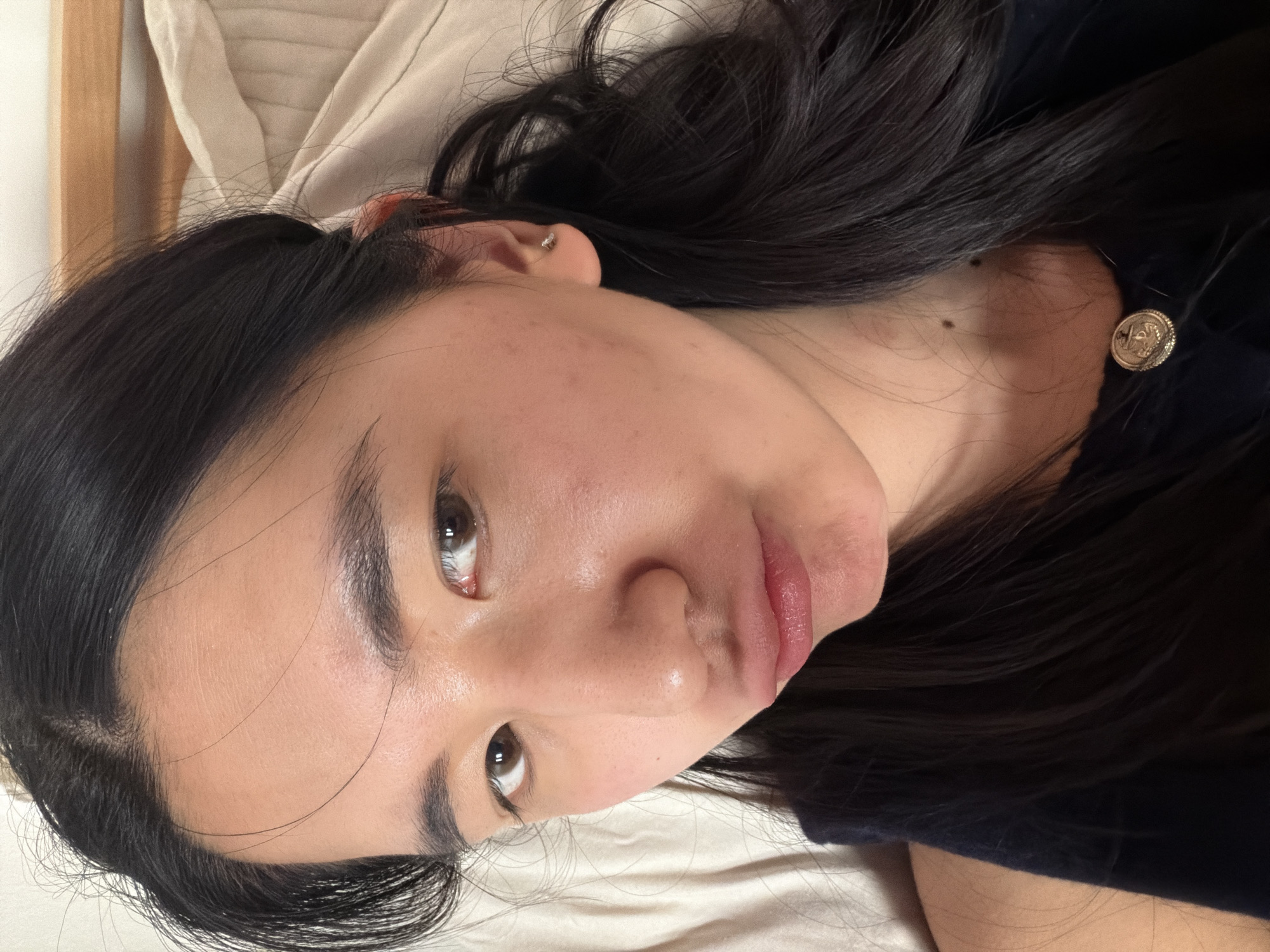 A close-up, angled shot of a woman's face. She has dark hair and is lying on a light-colored surface, possibly a bed. She has an ear piercing and a mole on her neck. A button is visible on her clothing. The background includes a cream-colored bed linen. There is no indication of a location in the city of Paris, France, in the image.