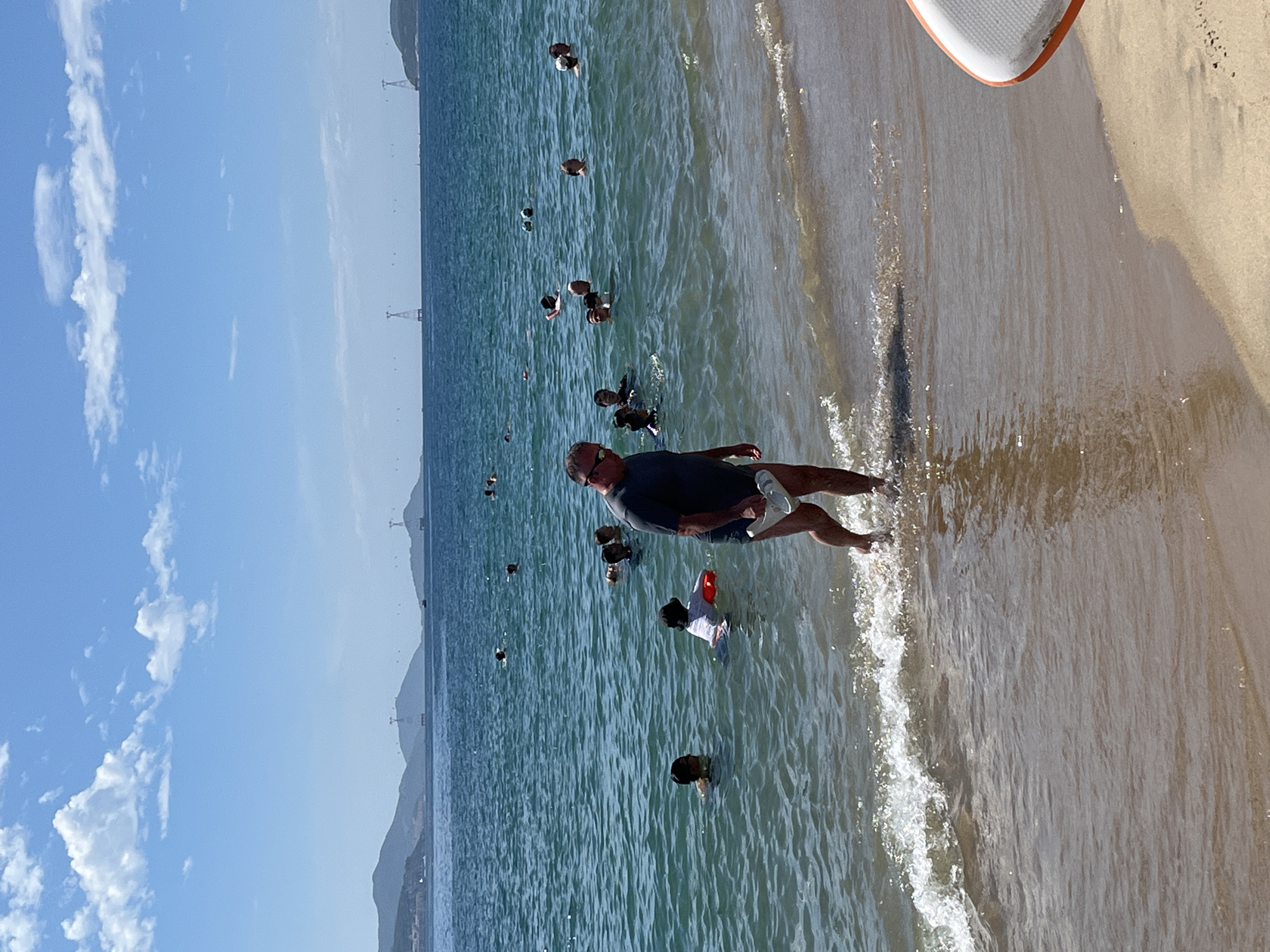 The image depicts a sunny day at a beach in Nha Trang, Vietnam. A man in a dark t-shirt and shorts walks out of the shallow water, holding a white object in his hand. He is on the right side of the frame, facing towards the left. In the background, several people are swimming and wading in the turquoise ocean. The water is calm with small waves lapping at the sandy shore. In the distance, mountains rise against a bright blue sky dotted with white clouds. Two tall tower structures with cables are visible on the horizon, likely part of a cable car system connecting to an island. A portion of an orange and white paddleboard is visible on the sand in the lower right corner.