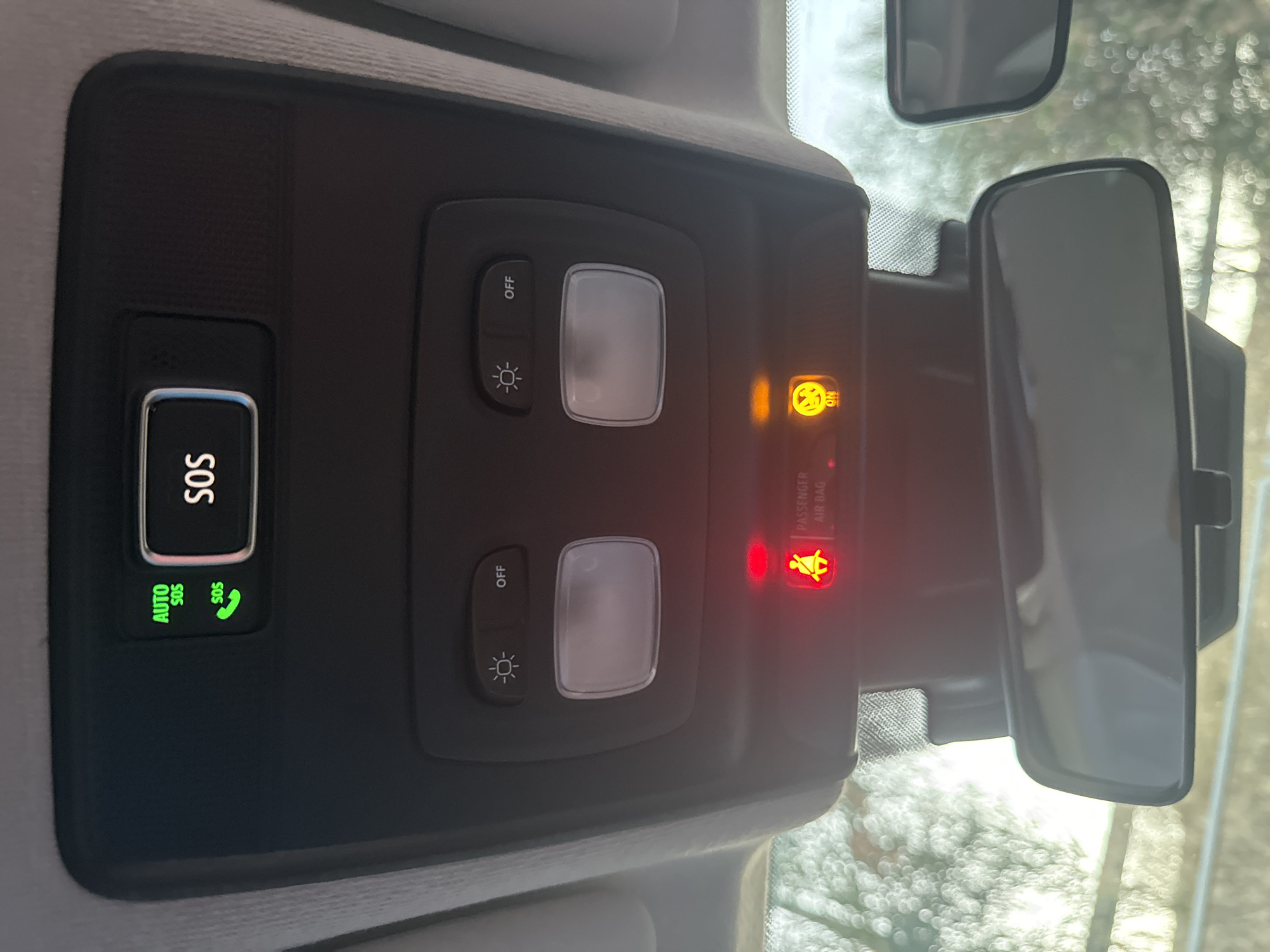 The image shows the interior of a car's ceiling console, featuring several buttons and lights. Prominently displayed is an SOS button with "AUTO SOS" indicated by green LEDs, alongside a green phone icon. To the right of the SOS button, the word "SOS" is illuminated in white text on a black background. Below this, there are two light controls, labeled with a sunburst icon and "OFF" respectively, each positioned above a translucent light fixture. Further down, two warning lights are visible: a red light with a seatbelt and "PASSENGER AIR BAG" icon, and an orange light with a child seat icon and the word "ON". A rearview mirror is centrally located below the console, reflecting a blurred outdoor scene. The image is taken from the perspective of someone inside the car, looking upwards. There is no information in the image to suggest a location in Murcia, Spain.