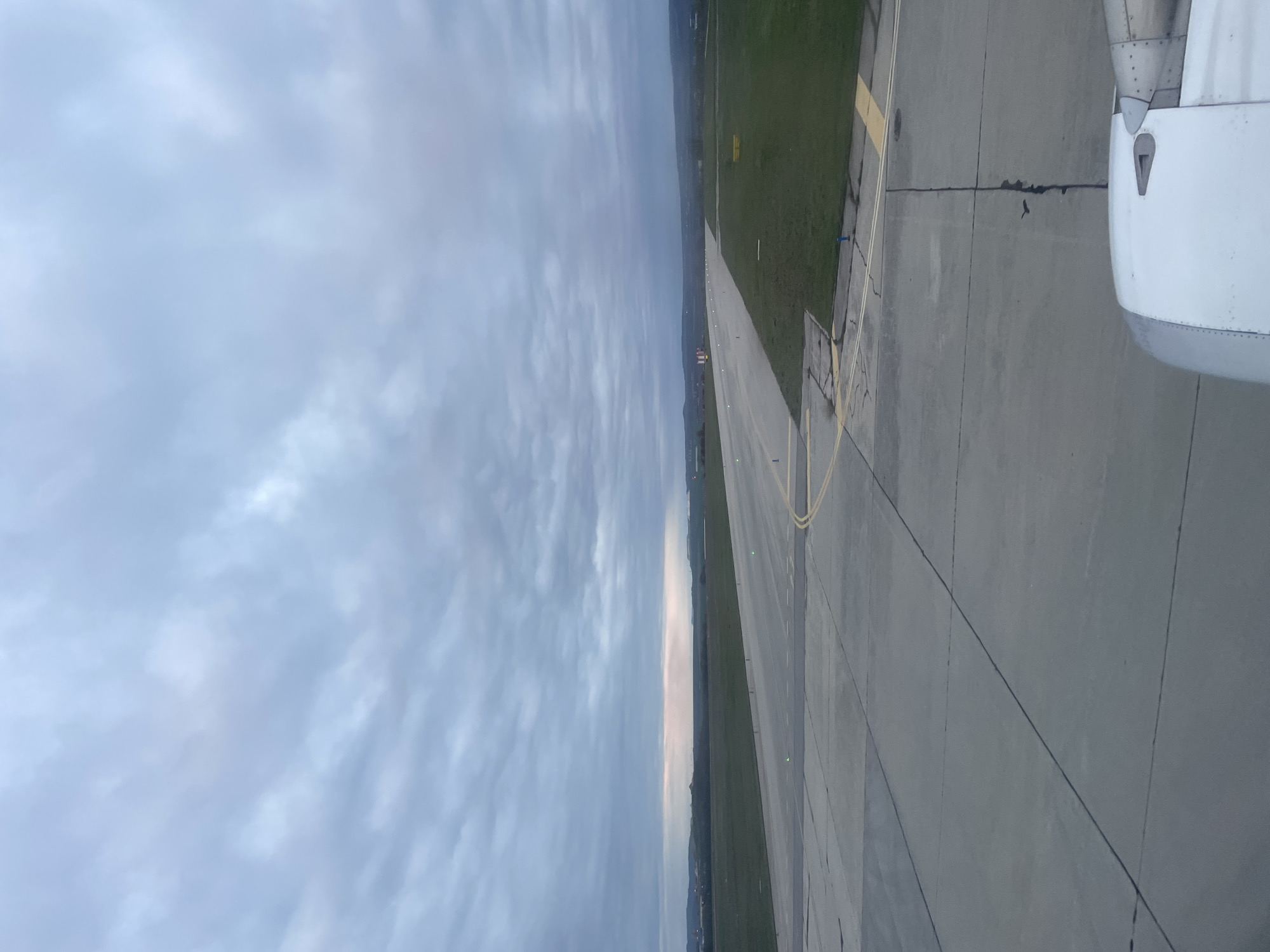 The image shows an airplane wing and engine positioned on a runway at an airport in Aksakovo, Bulgaria. The runway is gray and cracked, with yellow painted lines along the edges. Beyond the runway is green grass. The sky is mostly cloudy.