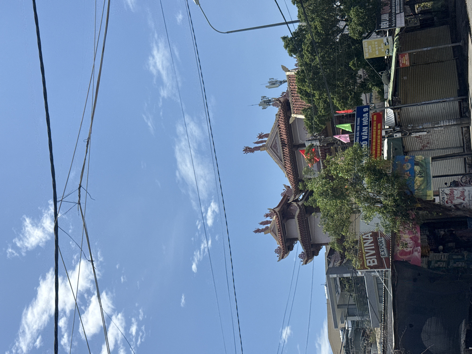 This is a daytime, eye-level, outdoor, full shot image of a street scene in Nha Trang, Vietnam, captured on a sunny day with a blue sky and scattered white clouds. In the foreground, power lines are strung across the frame, obscuring parts of the scene. A street lamp is visible on the right. In the midground, there are several structures, including a building with a traditional Asian tiled roof, adorned with decorative figures and cellular antennas on top. Adjacent to this are trees and various shops with closed metal shutters. Signs are visible, including "BIVINA EXPORT", "ĐỒ VĂN HÓA 9", and "BIỆN LẠNH". The overall impression is of a bustling commercial area with a mix of modern and traditional architecture.