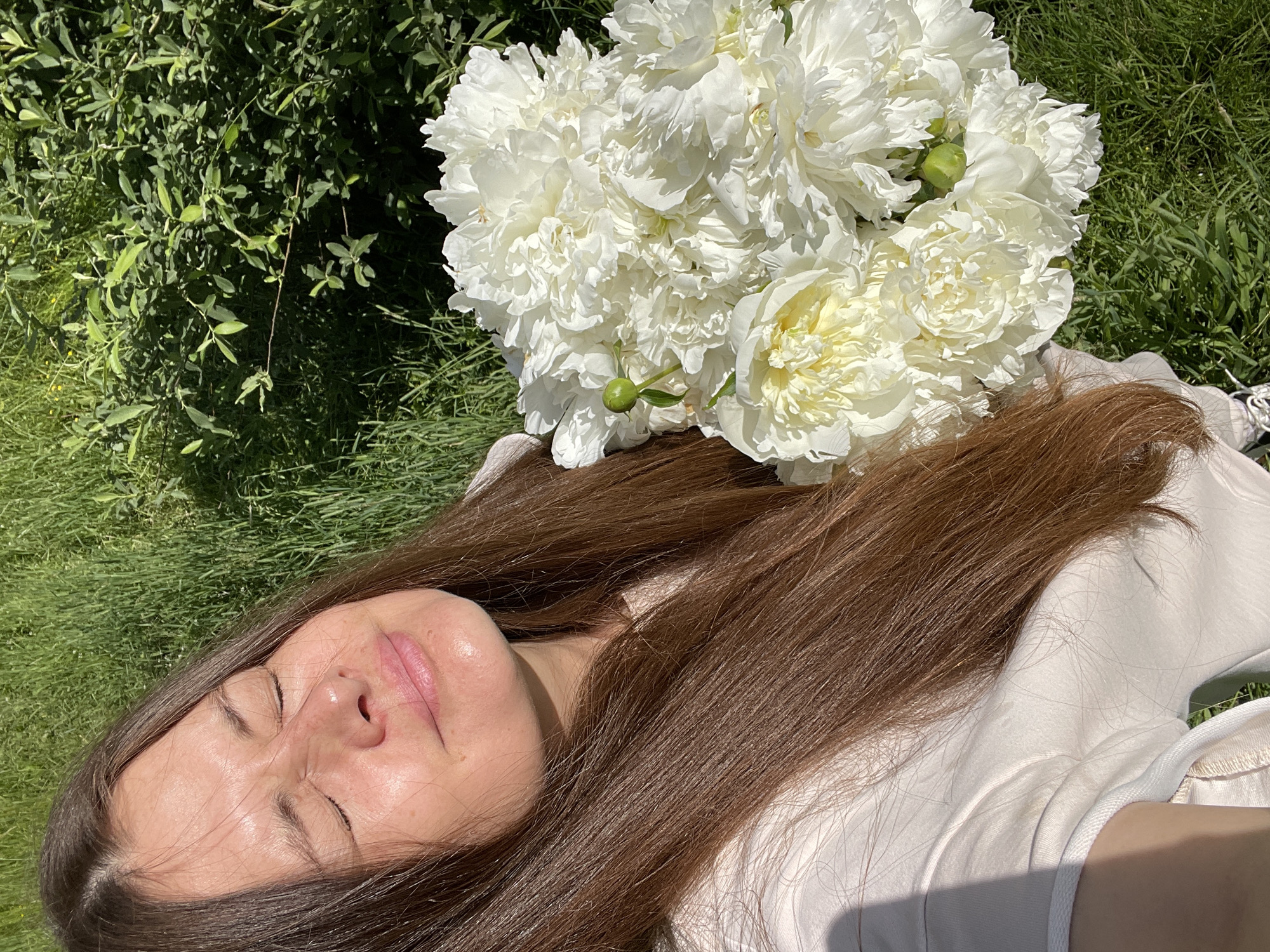 A woman with long, brown hair is holding a large bouquet of white peonies. She is outdoors in a grassy area, with green foliage in the background. The sun is shining, casting shadows on her face and clothing. Her eyes are closed, and her head is tilted back slightly, suggesting she is enjoying the moment. The peonies are fully bloomed, with many delicate petals. The scene appears to be a peaceful and natural setting.