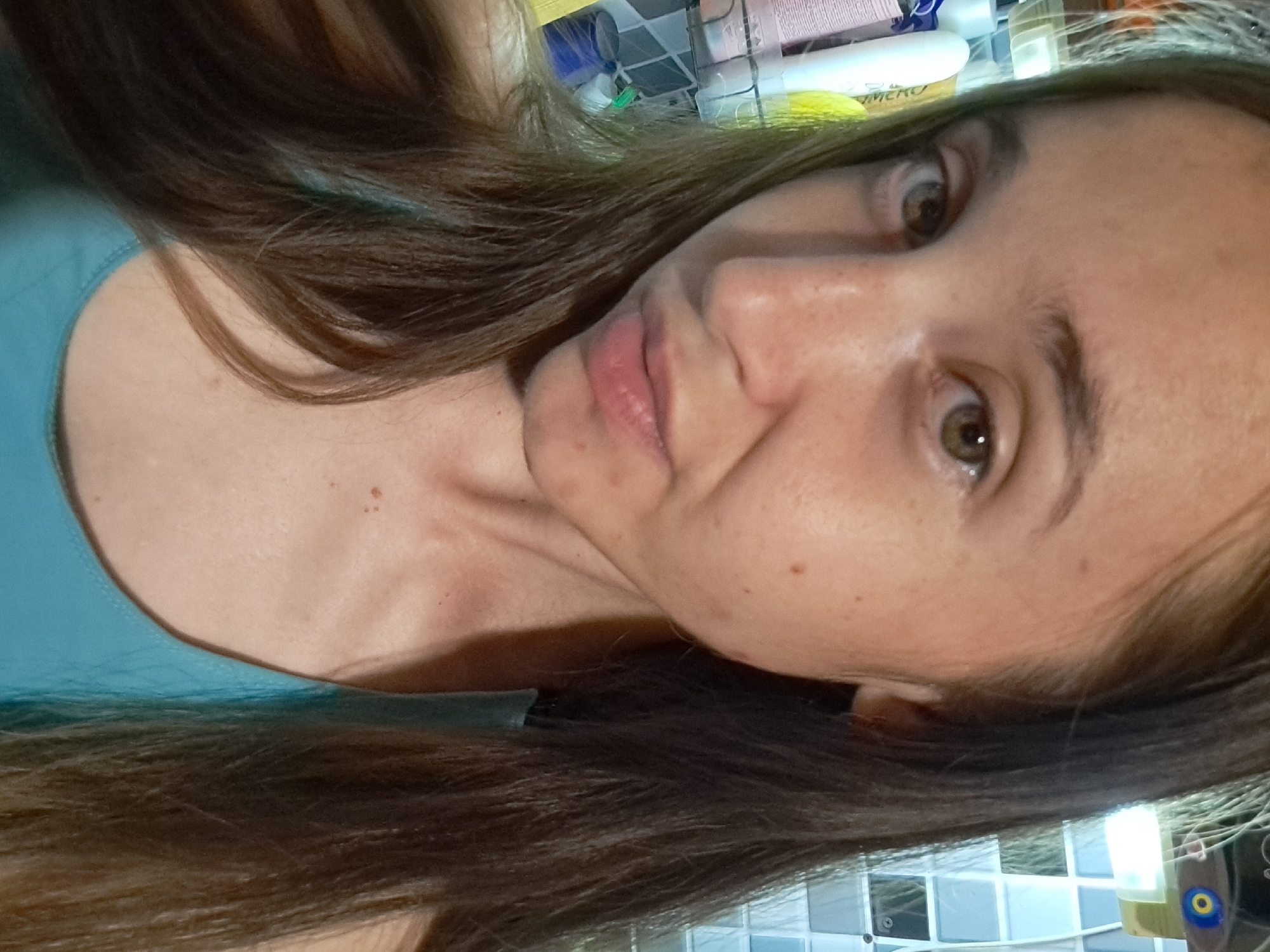 The image is a close-up, self-portrait of a young Caucasian woman. She has long, dark brown hair that frames her face, and her hair is slightly parted on the left. Her eyes are a hazel-green color, and her eyebrows are well-defined. Her skin has a fair complexion with visible freckles and a few blemishes on her cheeks and chin. She is wearing a teal-colored tank top, the straps of which are visible on her shoulders. Her lips are slightly parted, and her expression is neutral, with a slight tilt of her head. The background is blurred but appears to be a bathroom setting, with shelves stocked with various toiletries, including bottles and tubes of cosmetic products, and tiled walls. The lighting suggests an indoor environment, possibly illuminated by artificial light. The overall impression is that of a casual, personal moment captured by the subject herself. The image does not contain any specific information that would allow for its location to be identified within the city of Vypasne, Ukraine.