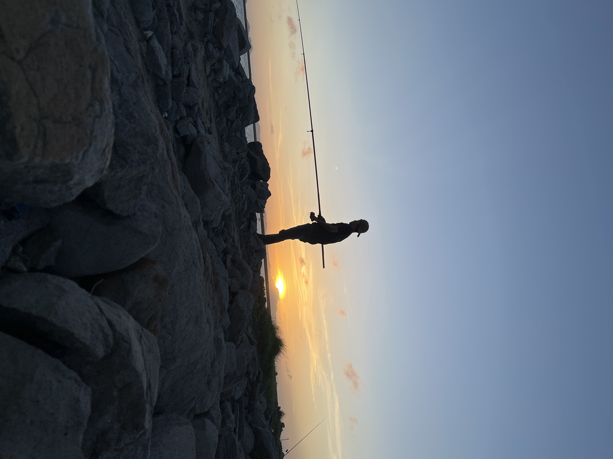 A silhouette of a lone fisherman stands on a rocky shore, casting his line into the water as the sun sets over the horizon. The sky is a gradient of soft blues and oranges, with a few wispy clouds tinged pink. The sea is calm, reflecting the colors of the sky. The fisherman, wearing a baseball cap and dark clothing, is the only human figure visible in the scene. The foreground is dominated by large, gray rocks of various sizes, creating a rugged texture. In the distance, faint outlines of hills or landmasses can be seen. The overall mood is serene and peaceful, capturing a moment of quiet solitude during sunset. This scene could be anywhere with a coastline, and without specific landmarks, it is not possible to confirm it is in Banda Aceh, Indonesia.