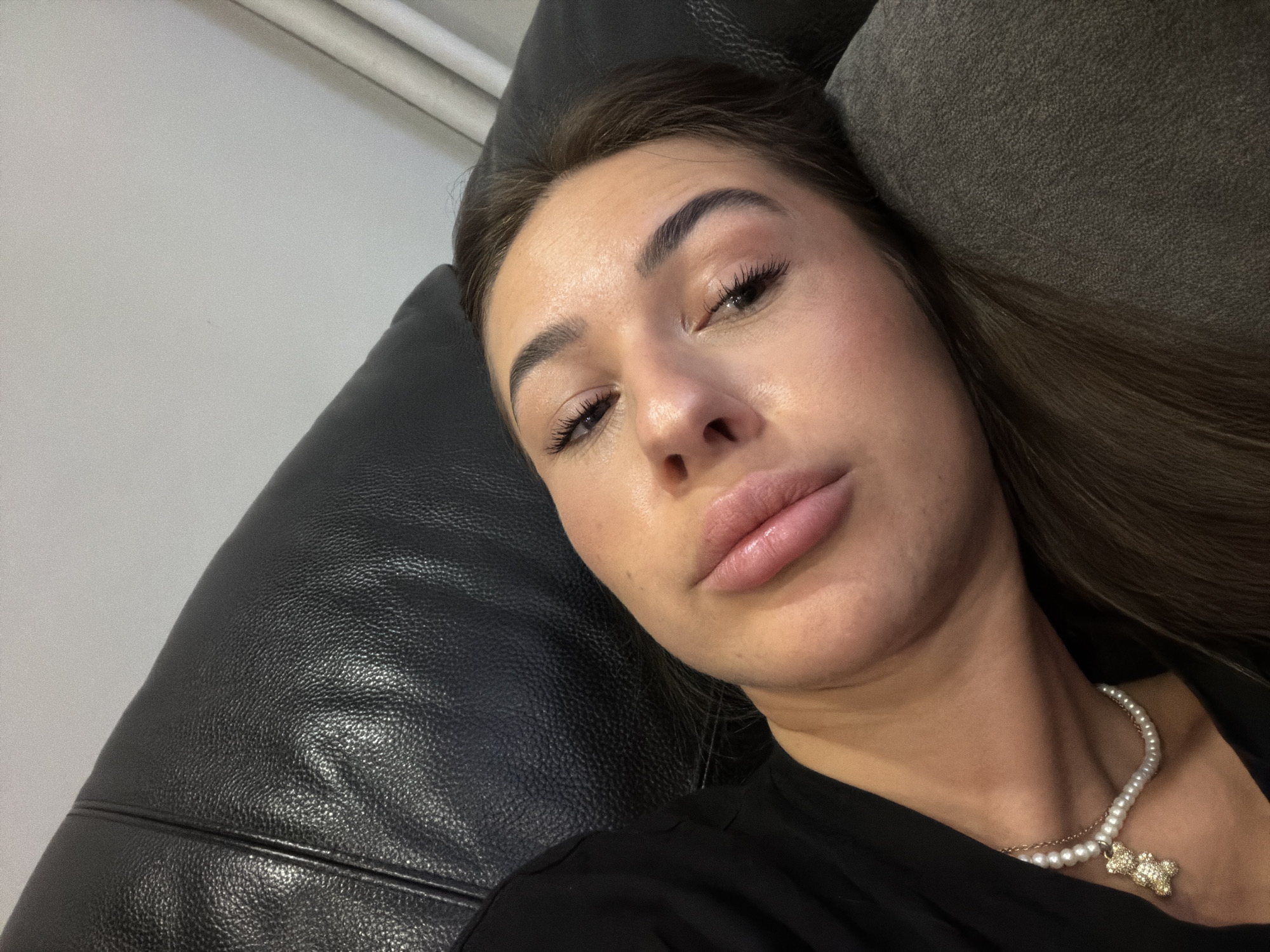 A young woman is lying down on a black leather couch in Novi Iskar, Bulgaria. She is wearing a black top and a pearl necklace with a teddy bear pendant. Her head is tilted to the side, and her eyes are closed. Her face is relaxed, with her lips slightly parted. The lighting in the room is soft, and there are some curtains in the background. The overall impression is one of peace and repose.