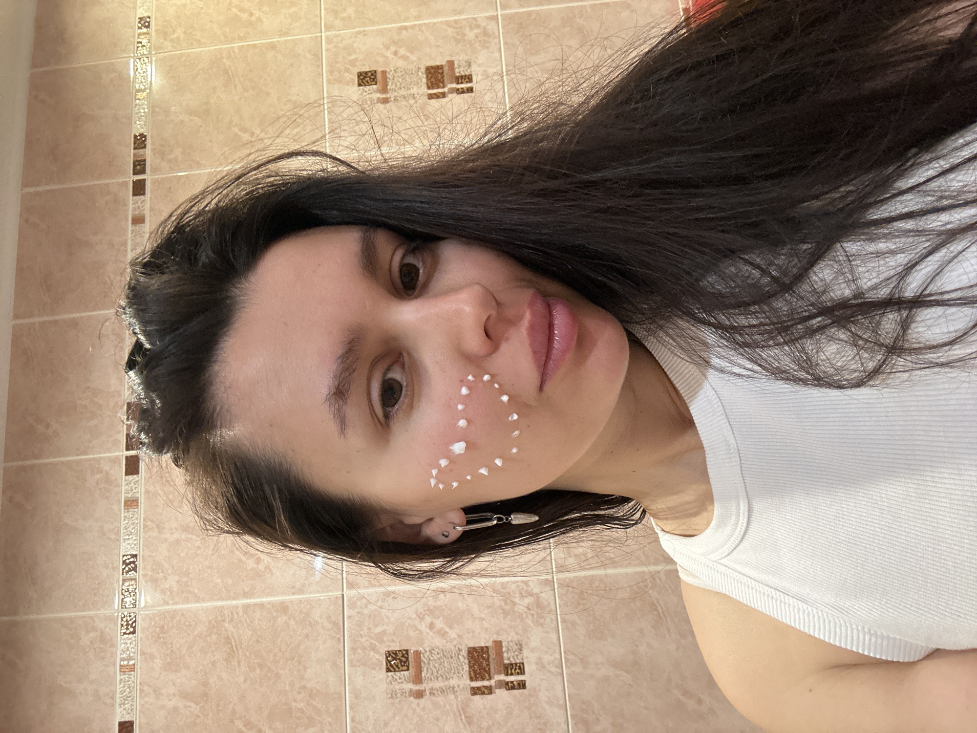 A young woman is facing the camera with white spots of cream on her right cheek. Her hair is dark and long, and she is wearing a white tank top. She is in a bathroom with tan and brown tiled walls. The location is not specified.