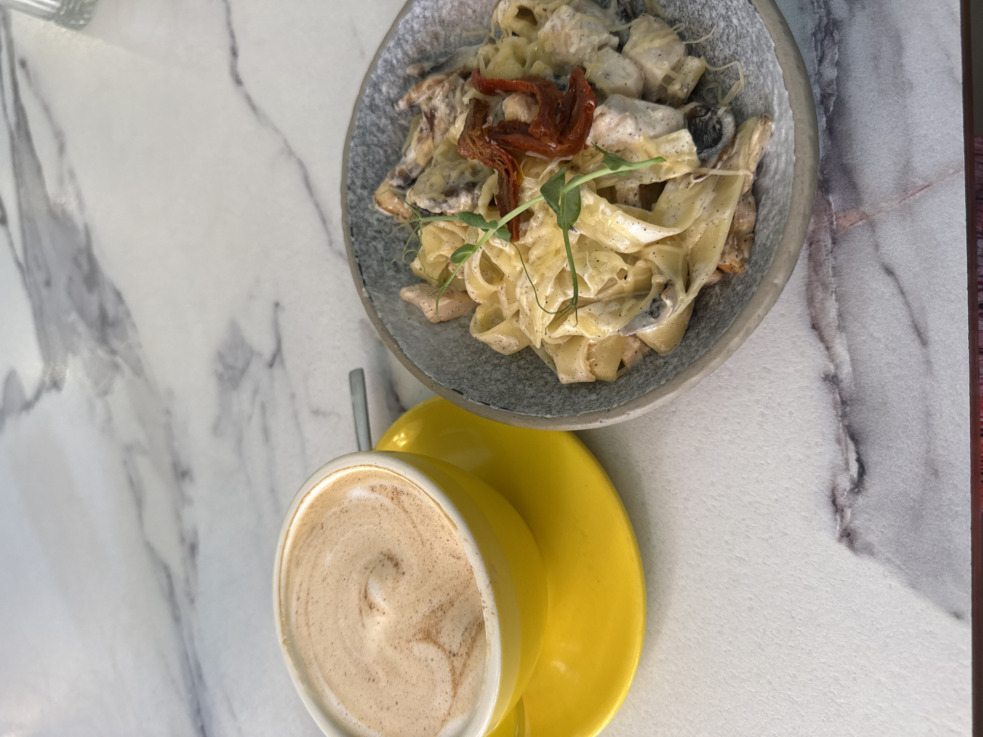An overhead, close-up shot features a yellow coffee cup filled with frothy coffee, positioned to the left on a yellow saucer. To the right, a gray bowl contains a serving of fettuccine alfredo with chicken and sun-dried tomatoes, garnished with a sprig of microgreens. The setting appears to be a cafe with a marble-patterned tabletop. This image does not contain any specific geographical markers that would allow for pinpointing its location within the city of Irkutsk, Russia.