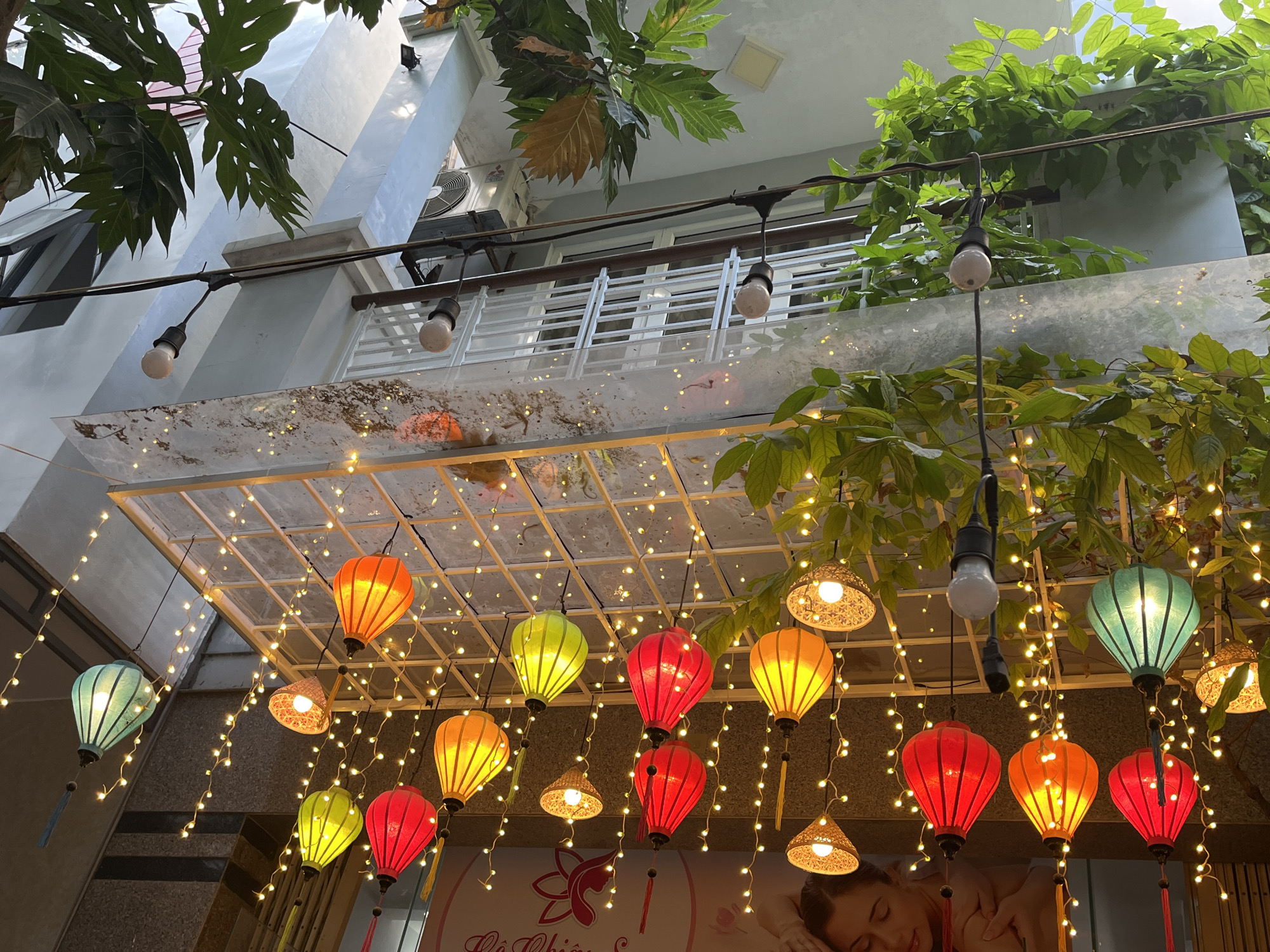 The image, captured from a low angle looking upwards, depicts a vibrant overhead display of colorful lanterns and string lights against the backdrop of a multi-story building in Đà Nẵng, Vietnam.
The main subjects are numerous traditional Vietnamese lanterns, varying in shape and color, including red, orange, yellow, green, and teal. These lanterns are illuminated from within, casting a warm glow. Interspersed among them are strands of small, warm white string lights, creating a sparkling canopy effect. Several exposed incandescent lightbulbs also hang from wires above the display.
Above the lighting fixtures, the facade of a light-colored building is visible. It features balconies with white railings and windows. Lush green foliage from trees and climbing plants frames the upper parts of the image, with some leaves partially obscuring the building and the sky. An air conditioning unit with "MITSUBISHI" visible on its casing is mounted on the building's exterior.
The glowing lights suggest the image was taken during the evening or night, creating an inviting and festive atmosphere. Part of a sign is visible at the very bottom, featuring a stylized logo resembling a lotus flower and partially visible text that appears to read "Lê Chiêu S..." (likely part of a business name). A woman's face, possibly from an advertisement for a spa or beauty service, is also faintly visible on the sign. The overall scene conveys a cheerful and decorative ambiance common in tourist areas or lively urban settings in Vietnam.