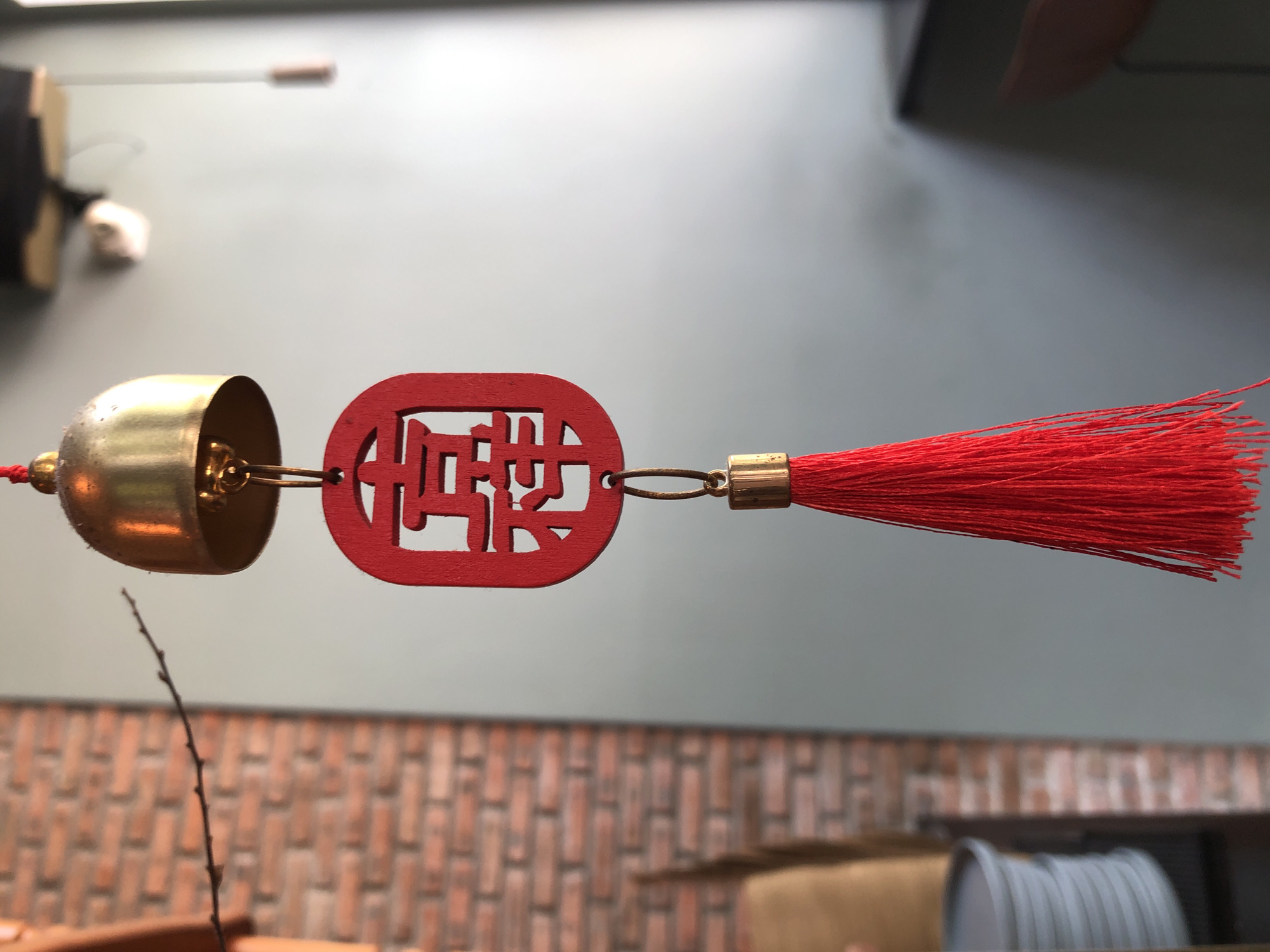 A close-up, eye-level shot focuses on a red Chinese character ornament hanging from a golden bell. The ornament, made of wood, features the characters "喜樂" which translate to "happiness and joy." Below the ornament, a vibrant red tassel dangles, adding a splash of color. The background is blurred, revealing a brick wall on the left and a gray wall on the right. The image appears to be taken indoors, possibly in a cafe or shop, as suggested by the presence of stacked plates in the lower left corner and a ceiling fan in the upper right. The lighting is natural, coming from a window on the right side of the frame. There are no people visible in the image. The setting is likely an establishment in Đà Nẵng, Vietnam, given the cultural context of the ornament and the general atmosphere of the scene.