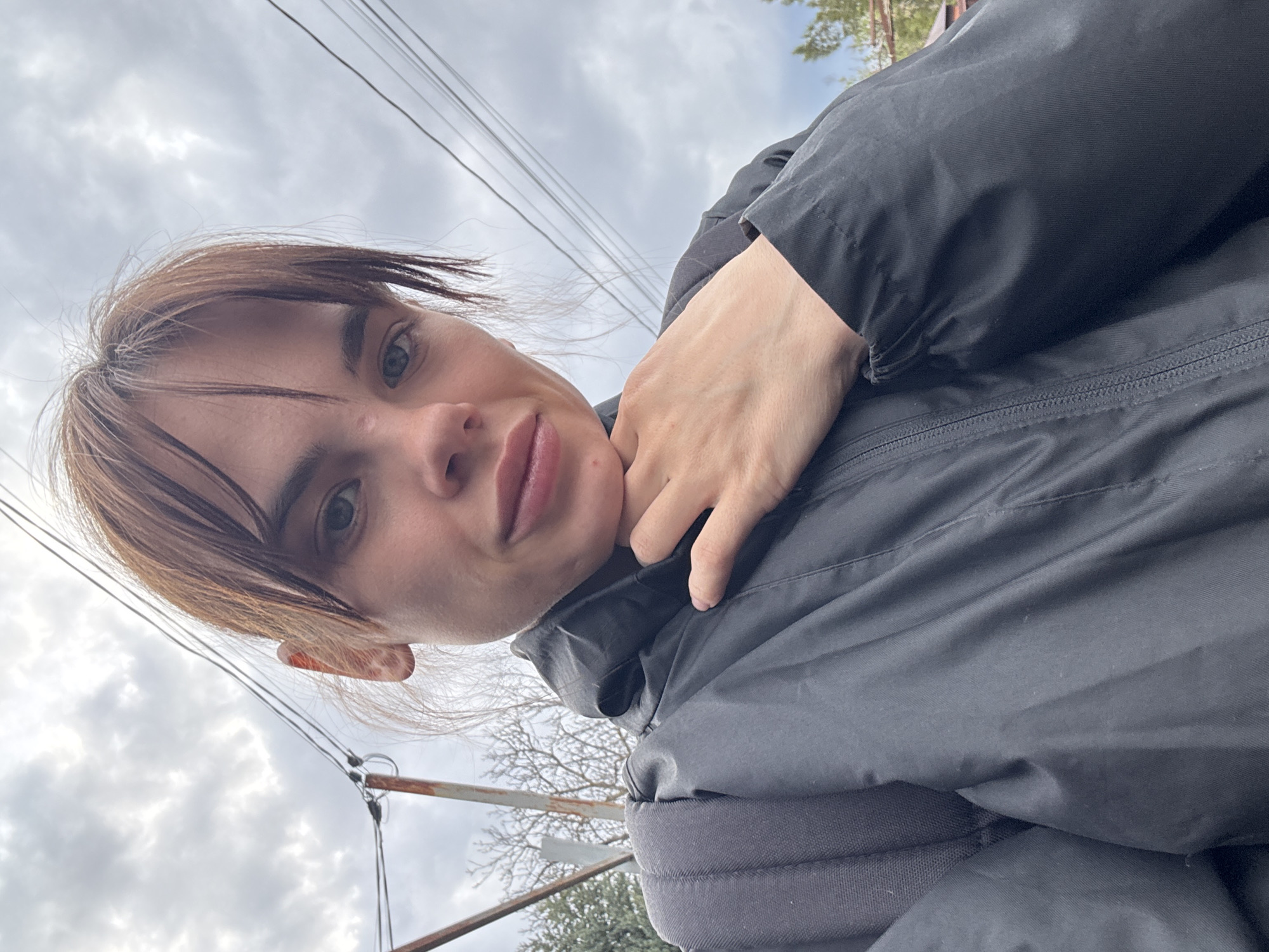 A person is posing for a selfie outdoors. They have fair skin, light brown hair with bangs, and are wearing a dark-colored jacket. The hand is resting on their chin. The background shows a cloudy sky, electrical wires, and glimpses of trees. Without additional information, it is impossible to determine if the picture was taken in Rostov-on-Don.