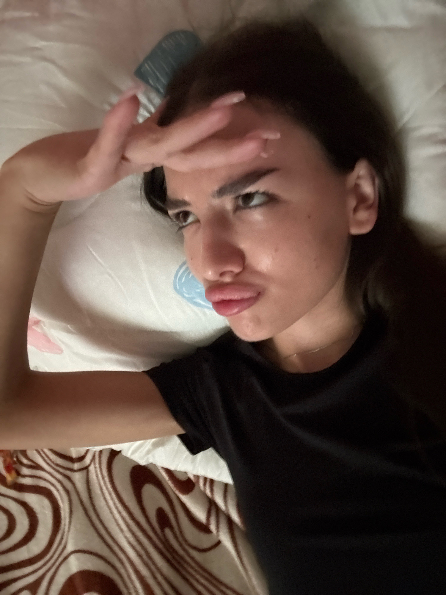 A young woman with dark hair is lying on a pillow and bed in what appears to be a bedroom. She is wearing a black t-shirt. Her left arm is raised, with her hand touching her forehead. Her lips are slightly pursed, and her eyes are open. The pillow she is lying on is white with a blue circular design. The bedsheets have a brown and white swirled pattern. The lighting is dim, suggesting it could be nighttime or the room is not well-lit. The image is a selfie, taken from a close-up perspective. No elements in the image suggest a specific location within the city of Basarabeasca, Moldova.