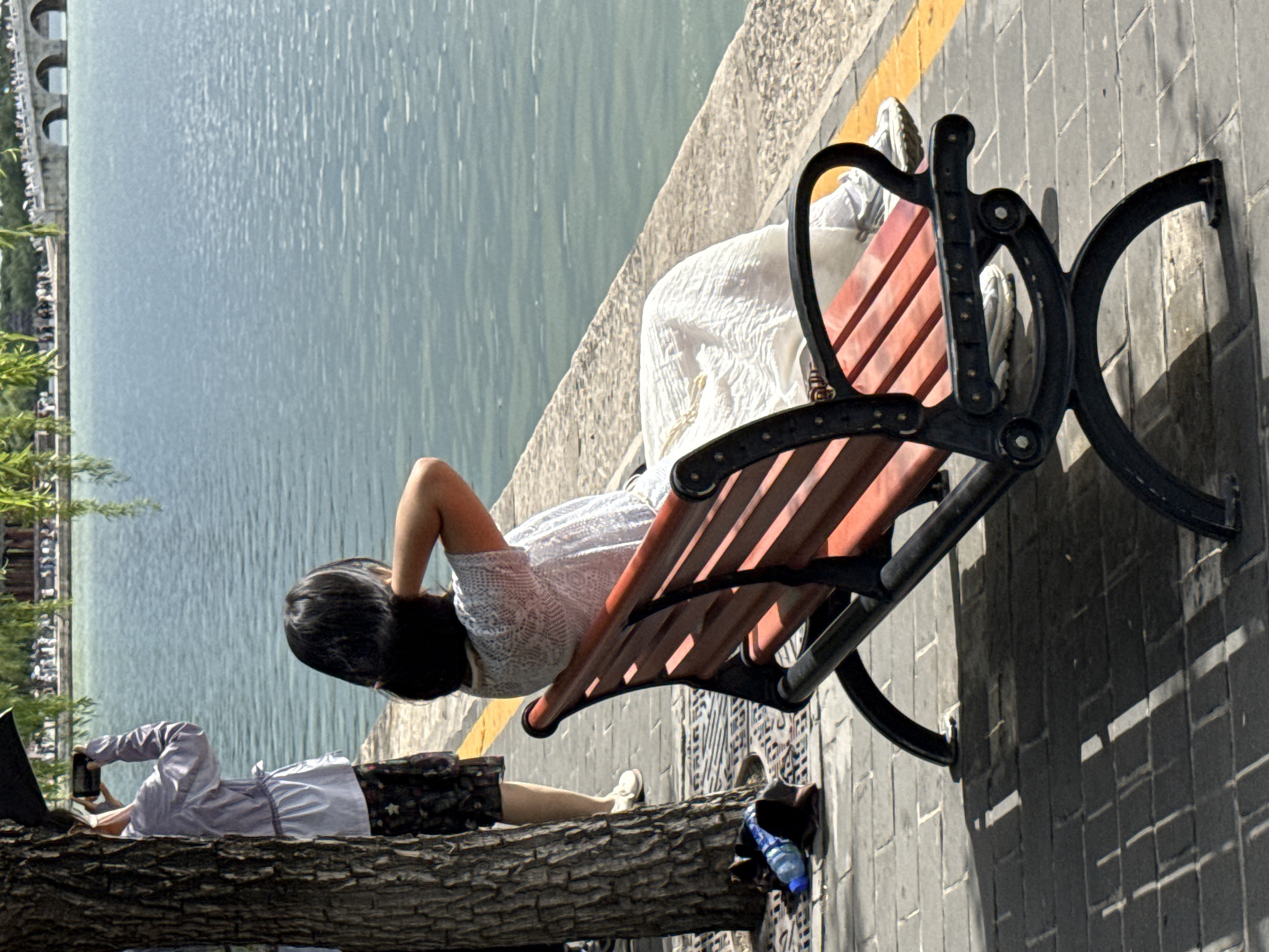 The image depicts an outdoor waterside scene in Haidian District, China, under bright sunlight. In the foreground, a woman is seated on a slatted reddish-brown wooden park bench with black metal supports, her back primarily to the viewer. She wears a light-colored, possibly white, patterned short-sleeved top and a flowing light-colored skirt, with light-colored athletic shoes on her feet. Her left arm is raised, with her hand positioned at the back of her dark hair. She is looking towards a large body of water.
To the left, partially obscured by a textured brown tree trunk, another person is standing. This individual wears a light-colored long-sleeved top cinched at the waist, dark patterned shorts or pants, and white shoes. They are holding a dark-colored smartphone horizontally, appearing to photograph or video the distant scene across the water.
On the paved grey brick walkway near the base of the tree, a blue plastic water bottle and a pair of dark slip-on shoes are visible. The walkway features drainage patterns and a distinct yellow line running along its edge. Beyond the walkway, a light-colored stone embankment meets the calm, light blue-green water, which exhibits surface ripples. In the far background, across the water, a multi-arched white or light-colored stone bridge is visible, with numerous small figures of people on it and along the distant shoreline. Green foliage, possibly from a weeping willow, hangs into the upper part of the frame. Strong sunlight casts sharp shadows of the bench and tree onto the paved ground.
