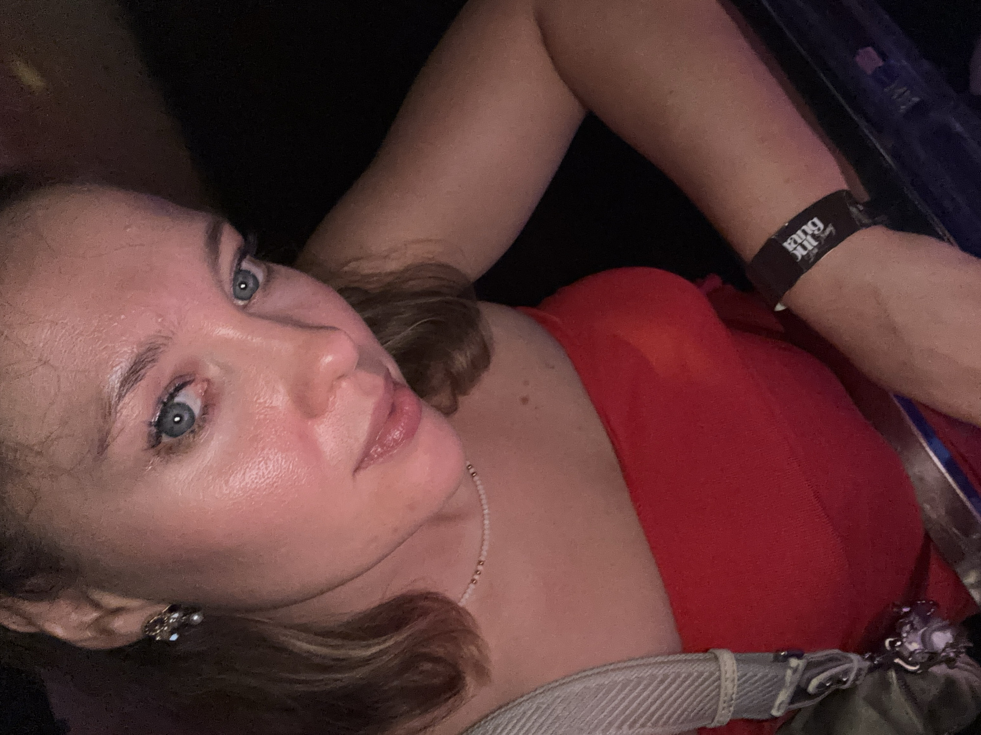 A close-up, high-angle selfie taken in a dimly lit environment. The subject is a young Caucasian woman with light skin, light brown hair pulled back, and blue eyes. She is wearing a red strapless top, a delicate beaded necklace, and a statement earring on her left ear. A light-colored purse strap is visible on her left shoulder. Her right arm is resting on a dark surface, and a black wristband with white text is visible on her wrist. The background is dark, obscuring details of the environment, but a hint of metal railing suggests a public venue, possibly a concert or event. The overall lighting is warm and intimate. There is no visible indication of the city of Đà Nẵng, Vietnam.