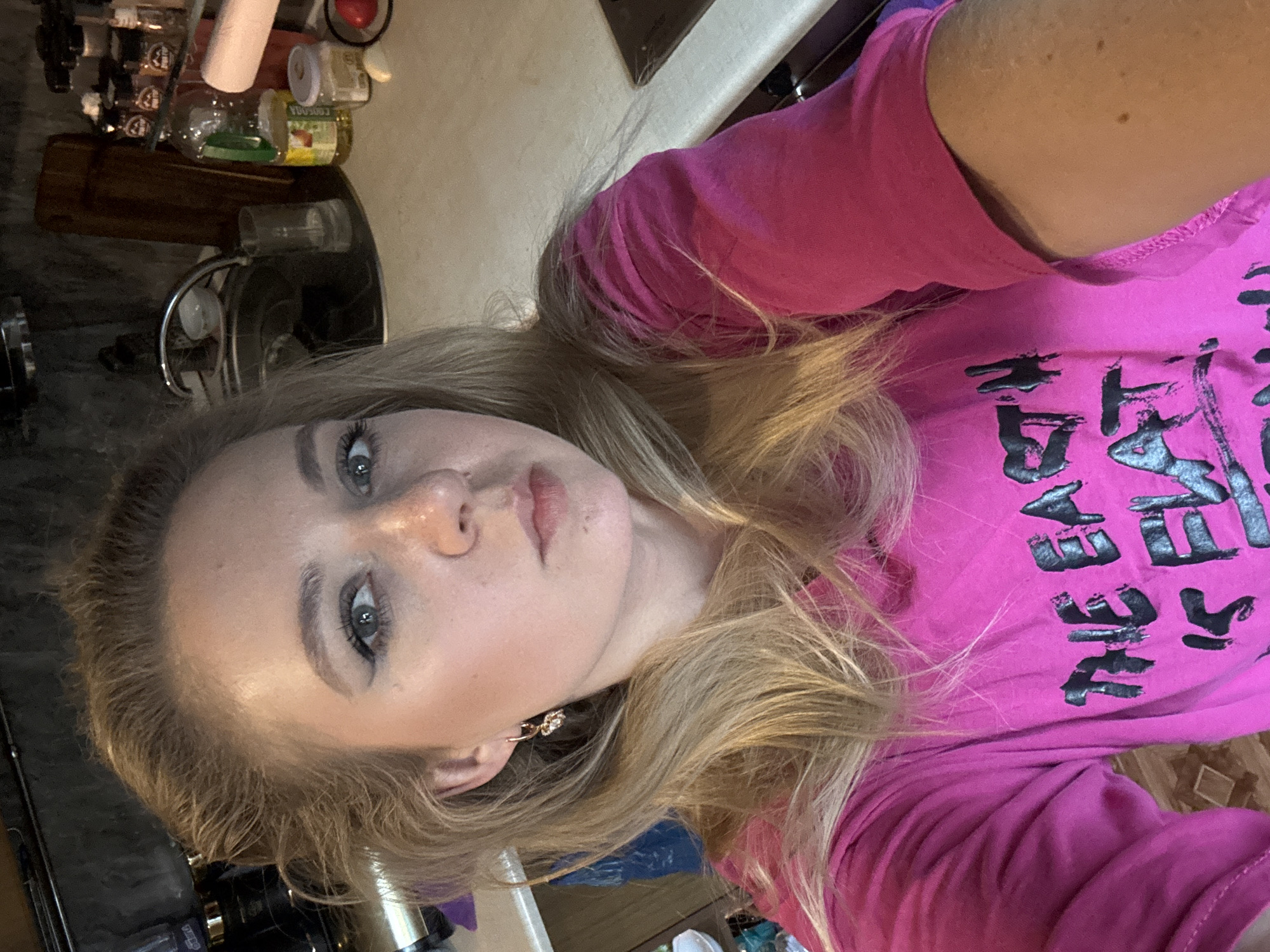 A woman with blonde hair and blue eyes is taking a selfie in what appears to be a kitchen. She is wearing a bright pink t-shirt with black lettering that says "THE EARTH IS FLAT". The background shows a kitchen sink, countertop with various items including spices, oil, and a piece of garlic, and a wooden cutting board. The lighting suggests an indoor setting, possibly during the day. The woman is looking directly at the camera.