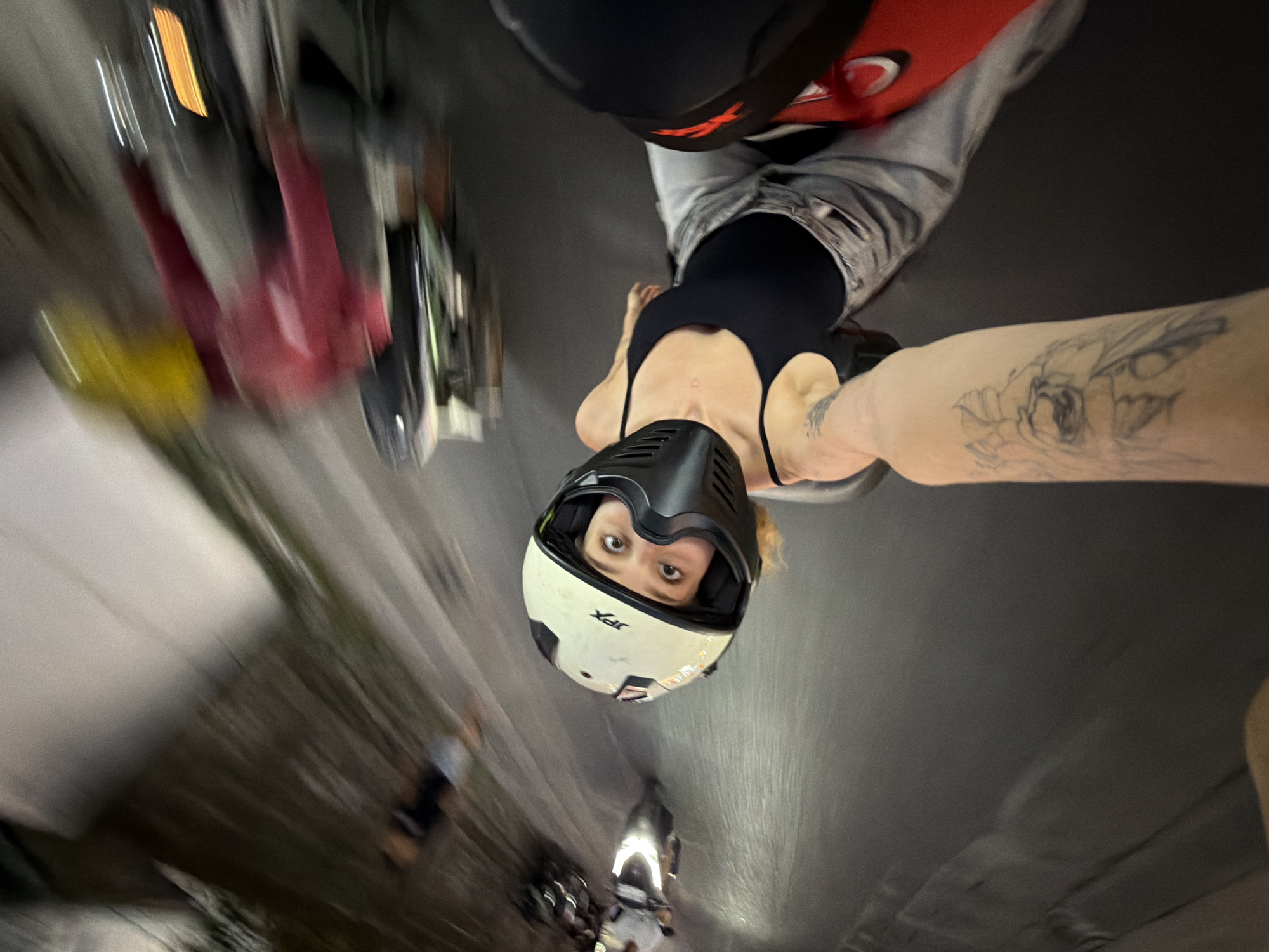 A selfie photo taken from the perspective of a person riding on a motorbike at night, likely in North Kuta, Indonesia, based on the visible context of multiple motorbikes, dense urban traffic, and distinctive street lighting. In the foreground, a person wearing a white and black full-face helmet with an open visor is looking directly at the camera. Their eyes are wide, and they have blonde hair. They are wearing a black tank top and grey pants, with a tattoo visible on their arm. The image is a motion blur, suggesting the motorbike is in motion. In the background, other motorbikes with riders are also visible, some with blurry figures in red and yellow attire. The road is dark with visible light streaks from headlights and streetlights. The overall impression is one of a dynamic, fast-paced nighttime scene on a busy Indonesian street.