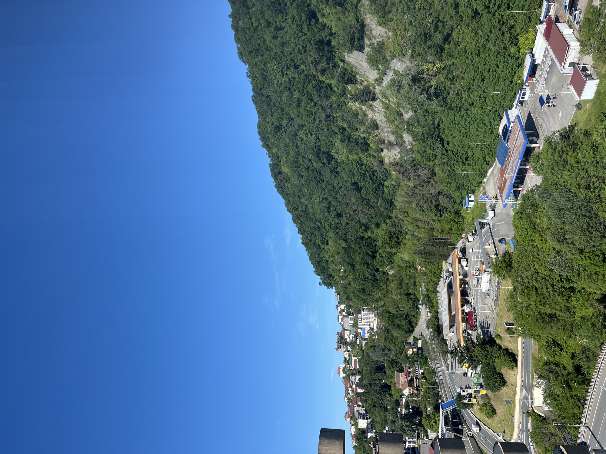 This panoramic image captures a vibrant daytime scene in a mountainous region, specifically Sochi, Russia, under a clear, expansive blue sky. The sky, devoid of clouds, dominates the upper left portion of the frame, indicating bright and sunny weather.
On the right side of the image, a large, steep mountain or hill rises dramatically, densely covered in lush green forestation. Some rocky outcrops are visible through the trees, highlighting the rugged terrain.
Below the forested mountain, the landscape transitions into a developed valley area featuring a network of roads, buildings, and human activity. A significant multi-lane road winds through the scene, populated by various cars and trucks. A prominent feature in the mid-ground is a large gas station or rest stop complex, identifiable by its distinct canopies (one blue, one with an orange/brown underside) and multiple fuel pumps. Several vehicles are parked or actively using the facilities.
Further along the roads and nestled into the lower parts of the hillside, numerous buildings are visible, including residential-style houses with varying roof colors, suggesting a thriving community or settlement.
In the immediate foreground, sturdy, curved concrete structures are partially visible, indicating that the photograph was taken from an elevated vantage point, possibly from a bridge, an overpass, or a high-rise structure overlooking the valley.
The text "ЛУКОЙЛ" (Lukoil), a Russian oil company, is clearly visible in white letters on the blue canopy of the gas station, serving as a specific location cue. The scene portrays a blend of natural beauty and functional urban infrastructure.