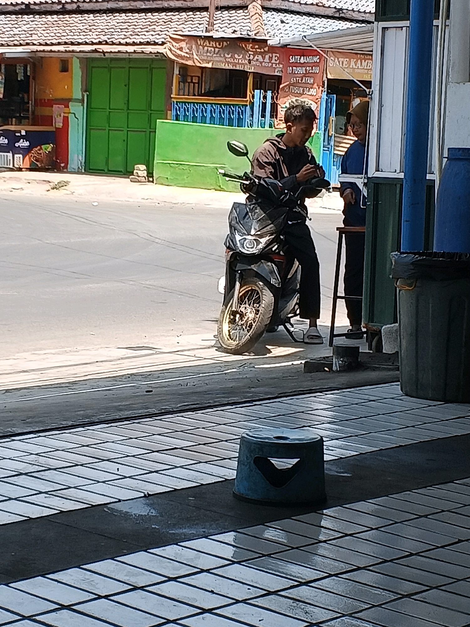 In Taman, Indonesia, a man in a brown jacket and black pants is seated on a motorcycle. He appears to be looking at his phone or engaging in some activity with his hands. To his right, a person wearing a blue shirt and headscarf stands behind a counter, possibly interacting with the man. Behind them, a building with colorful walls and a green gate is visible. The scene appears to be near a road, with a motorcycle parked on the side. The ground in the foreground is tiled.
