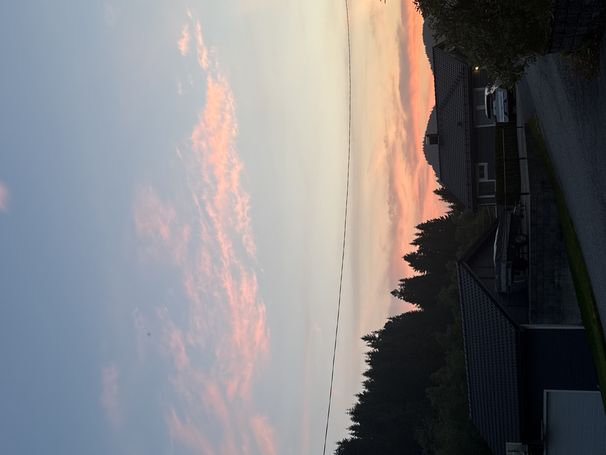 A low-angle, vertical, medium shot captures a serene sunset over a residential area in Valestrandsfossen, Norway. The sky is a canvas of soft blues and pinks, with wispy clouds stretching across the upper half of the frame. A thin black wire traverses the middle of the sky horizontally, suggesting overhead power lines. Below the sky, a dense line of dark evergreen trees forms a silhouette against the colorful sunset. In the foreground, two modern houses are visible. The house on the left has a dark gray tiled roof and a dark blue siding, with a white garage door. A trailer is parked to the right of this house, resting on a low brick wall. The house on the right is also dark gray with a tiled roof, and a white SUV is parked in its driveway. The road leading to the houses is paved and curves gently through the scene. The overall atmosphere is peaceful and quiet, indicative of dusk.