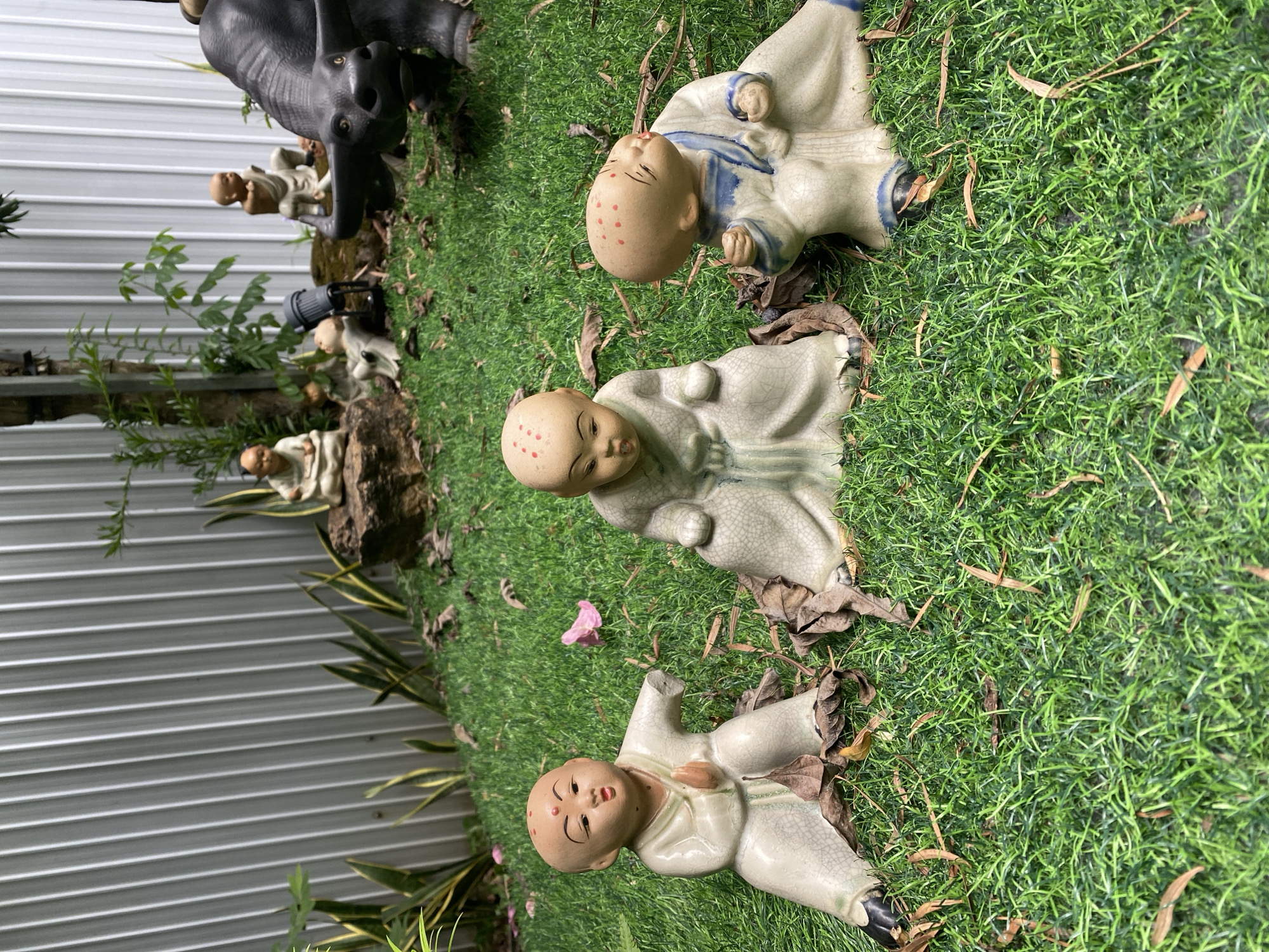 A group of ceramic figurines, appearing to be young Buddhist monks, are displayed on a patch of green artificial grass. Three prominent figurines are in the foreground, each with red dots on their shaved heads and wearing traditional robes. The figurine on the left is in a seated, praying posture, while the middle figurine is also seated, looking forward. The figurine on the right is leaning back with its eyes closed and a smile. Behind these, several other similar figurines are visible, some seated on rocks and others in the background. A large, dark-colored bull figurine is positioned to the right and slightly behind the foreground monks. The background consists of a corrugated metal fence and some sparse vegetation. The scene suggests an outdoor decorative display, possibly in a garden or courtyard. The image appears to be taken in Da Nang, Vietnam, as the location is specified.