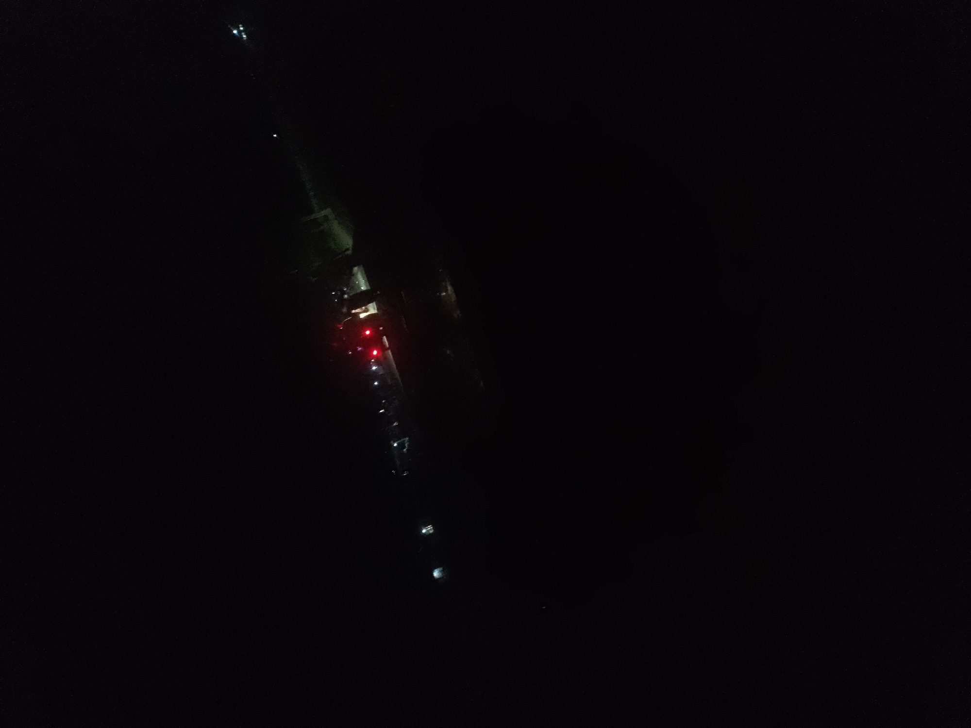 The image is a dark, aerial view of a road or pathway at night in Jalingo, Nigeria. Several red lights are visible, likely from vehicle taillights, indicating traffic or a stationary line of vehicles. Some white lights are also present, possibly from headlights or streetlights, but they are less distinct. The surrounding environment is predominantly black, suggesting a lack of ambient light and possibly a rural or less developed area. The image is blurry and grainy, making it difficult to discern specific details about the vehicles or the surroundings. The perspective suggests the photo was taken from a high vantage point, such as a drone or a tall building.