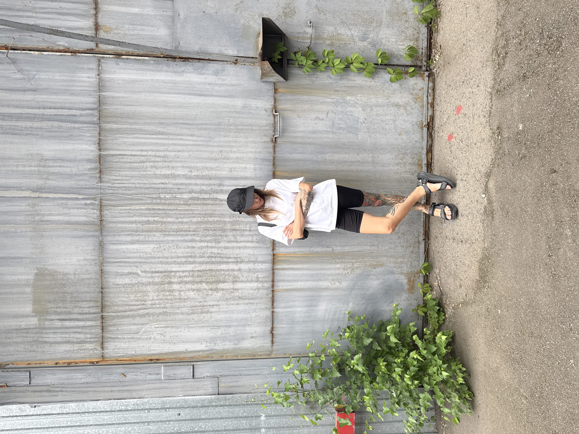 A person with long blonde hair and a black baseball cap stands with their arms crossed in front of a corrugated metal wall. They are wearing a white t-shirt and black shorts, and have tattoos on their arms and legs. The person is wearing black sandals and is positioned in front of a large, weathered metal door. Green vines with heart-shaped leaves are growing up the wall, emerging from a metal chute near the top of the door.
The ground next to the wall is paved with asphalt, with two small red markings visible. The scene appears to be outdoors, possibly in an industrial or urban setting. The lighting suggests it is daytime, and the weather seems overcast or diffused. There is no visible text in the image. The overall mood is casual and relaxed.
