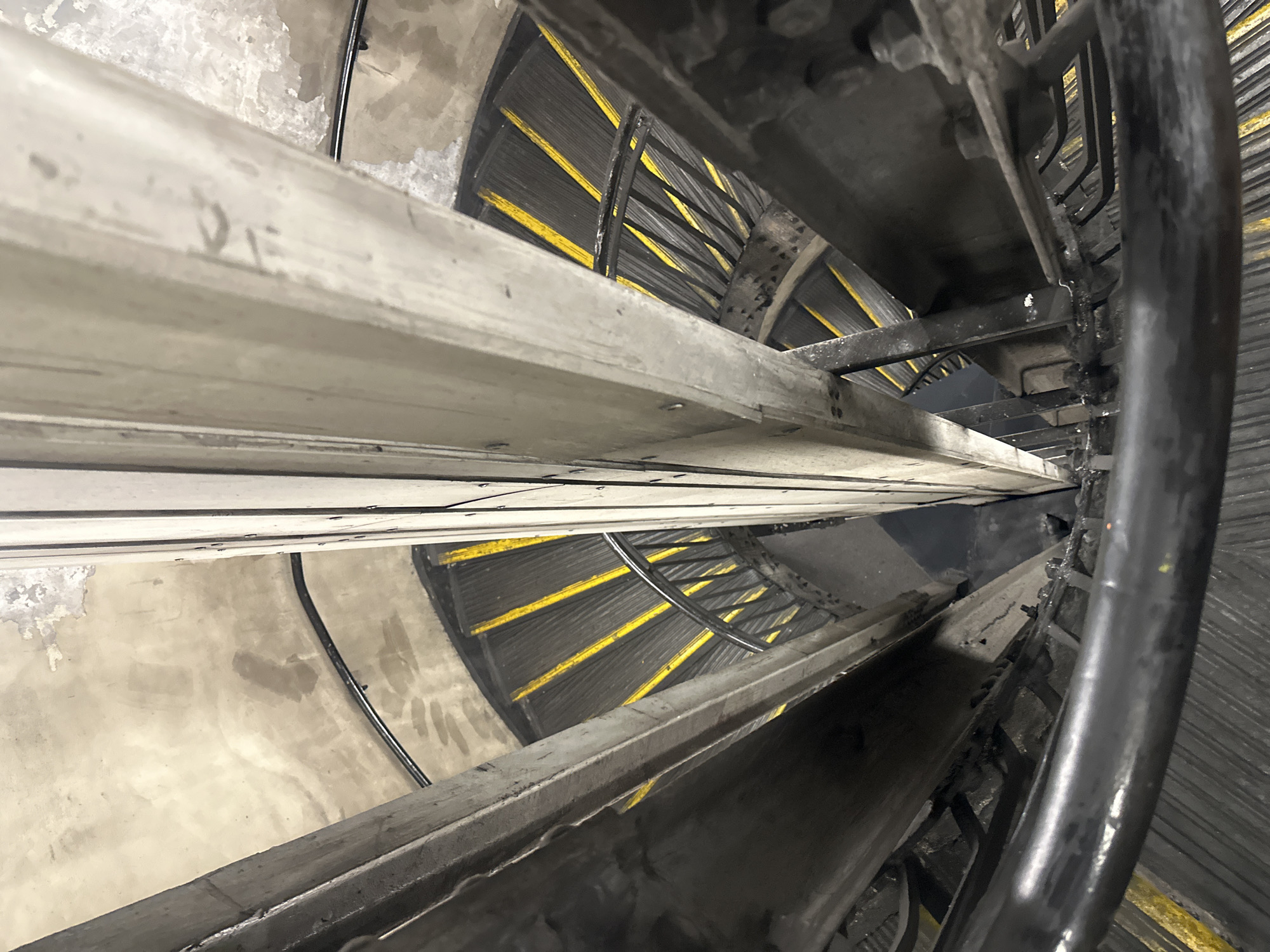 The image depicts the interior of a spiral staircase, likely within a commercial or industrial setting due to the presence of cable trays and other metallic structures. The staircase is constructed of metal and has a handrail on one side. The steps are lined with black rubber or plastic strips that have yellow accents. The walls are rough and appear to be made of concrete. The environment seems enclosed and dimly lit. There are no people visible in the frame. The location of this image is the British Telecom (BT) Tower in London, United Kingdom.