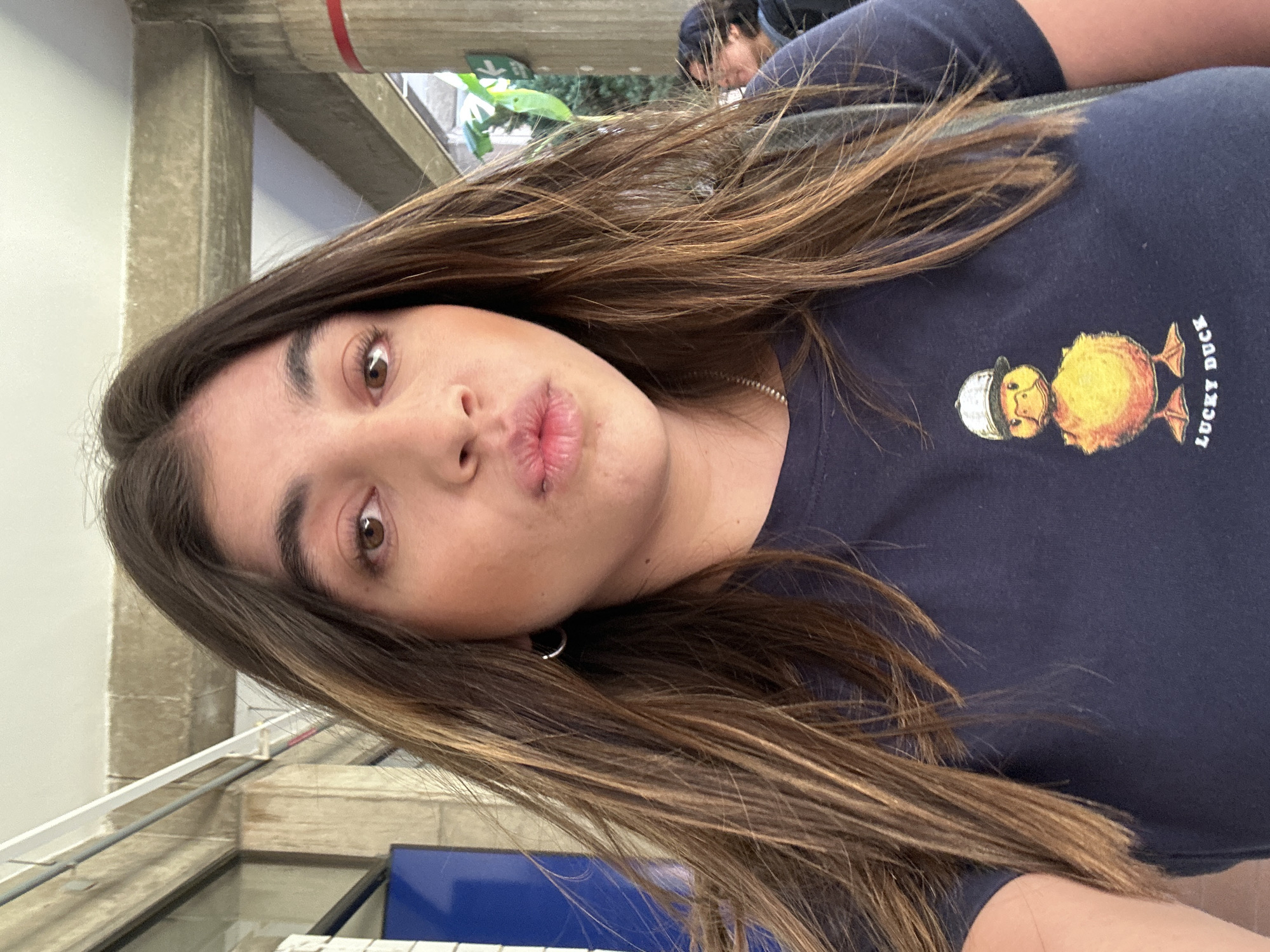 An image captured in San Bernardino Tlaxcalancingo, Mexico, presents a close-up, waist-up view of a young female subject. She possesses long, dark brown hair with lighter highlights, parted centrally, and brown eyes. Her lips are pursed into a puckered expression. She is wearing a dark blue short-sleeved t-shirt featuring a graphic of a yellow duck wearing a white baseball cap, with the words "LUCKY DUCK" printed below it in white text. A thin silver-colored chain is visible around her neck, and a small silver hoop earring is present in her left ear. The background shows structural elements consistent with an urban or institutional setting. Visible features include a segment of a blue rectangular object to the left, likely part of a door or frame, and overhead concrete beams. A white horizontal conduit or pipe runs along the upper left. To the right, a concrete pillar with a red horizontal stripe is partially visible, behind which green foliage suggests outdoor plant life. A blurred figure of another individual with dark hair is partially discernible in the far right background. The scene is illuminated by daylight.