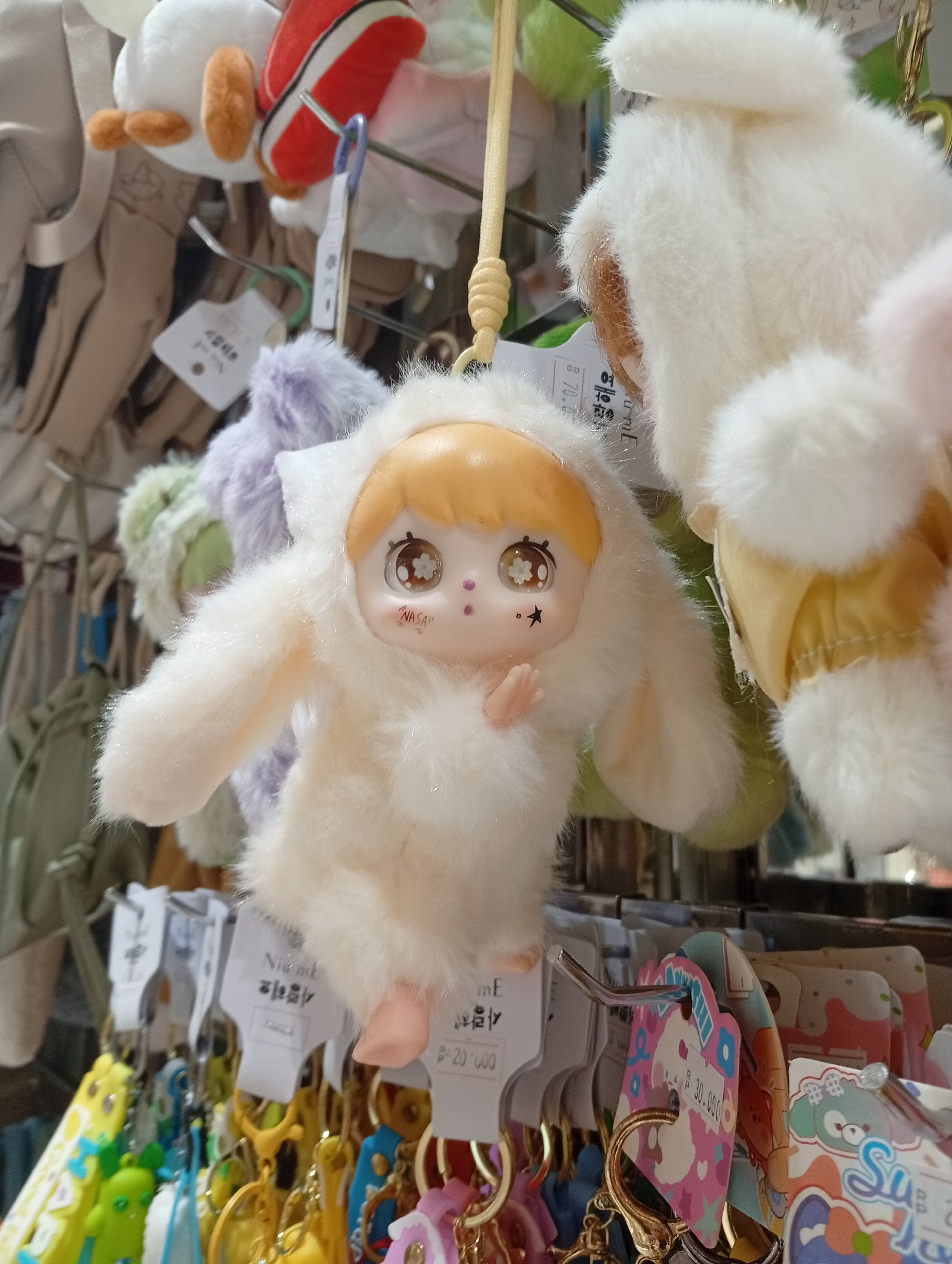 The image shows a close-up of a keychain display, likely inside a shop. A plush doll with an anime-style face and a furry white costume is the main focus. The doll has blonde hair, large eyes with star details, and the word "NASA" printed on its cheek. It has one hand raised. It hangs from a golden-colored keychain. Behind it are other plush keychains in different colors and styles. A metal rack holds the keychains, and price tags in Korean are attached beneath each. Below the keychains, there is a blur of colorful keychain designs. Some of the visible price tags mention the price as "B 70.00" and "B- 20 000" and "300.000". This is likely in Denpasar, Indonesia.