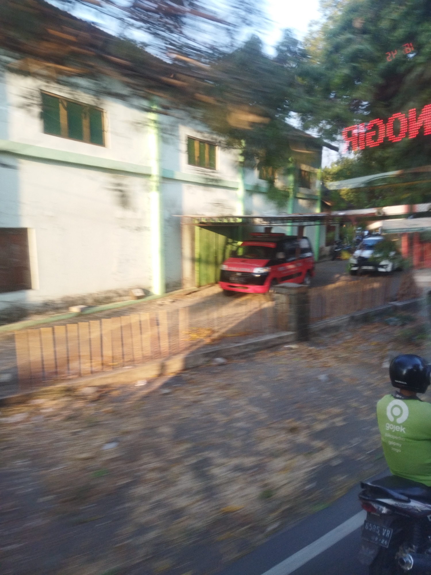 The image captures a street scene with motion blur, suggesting movement, possibly from a vehicle. A light-colored building with green-framed windows is visible on the left. In front of the building, a covered parking area shelters a red and black van. To the right of the van, a white car is parked. Further to the right, a person wearing a green Gojek jacket and a black helmet is riding a motorcycle. The license plate on the motorcycle reads "6595 VR". Red LED lights displaying "ROOM" are visible on a surface reflecting the light. The ground is covered with fallen leaves and debris. The overall environment appears to be an urban or semi-urban setting in Singodutan, Indonesia.