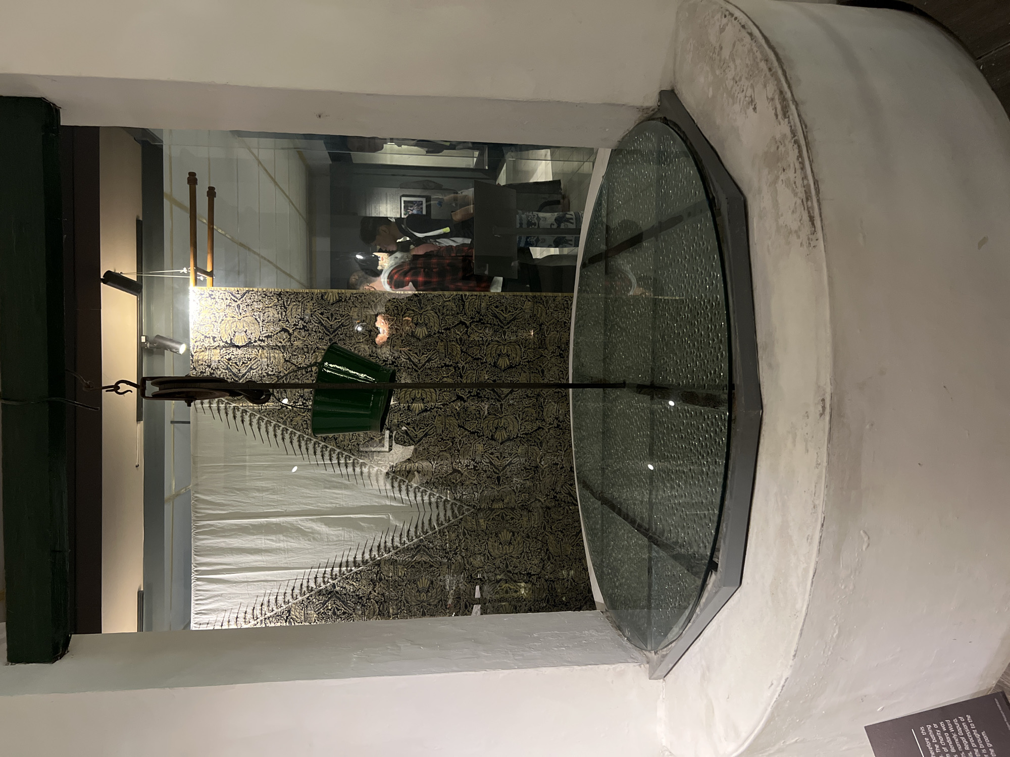The image shows a well with a glass cover in a museum in Yogyakarta, Indonesia. The well appears to be a historical artifact, possibly part of a traditional house or a public space. A green bucket is suspended by a rope from a pulley system above the well, suggesting it was used for drawing water. The surrounding area is decorated with traditional Indonesian textiles featuring intricate patterns. In the background, there are visitors in the museum, some of whom are looking at exhibits. The well is situated within a building, indicated by the interior lighting and the presence of museum displays. The overall scene conveys a sense of historical preservation and cultural exhibition.