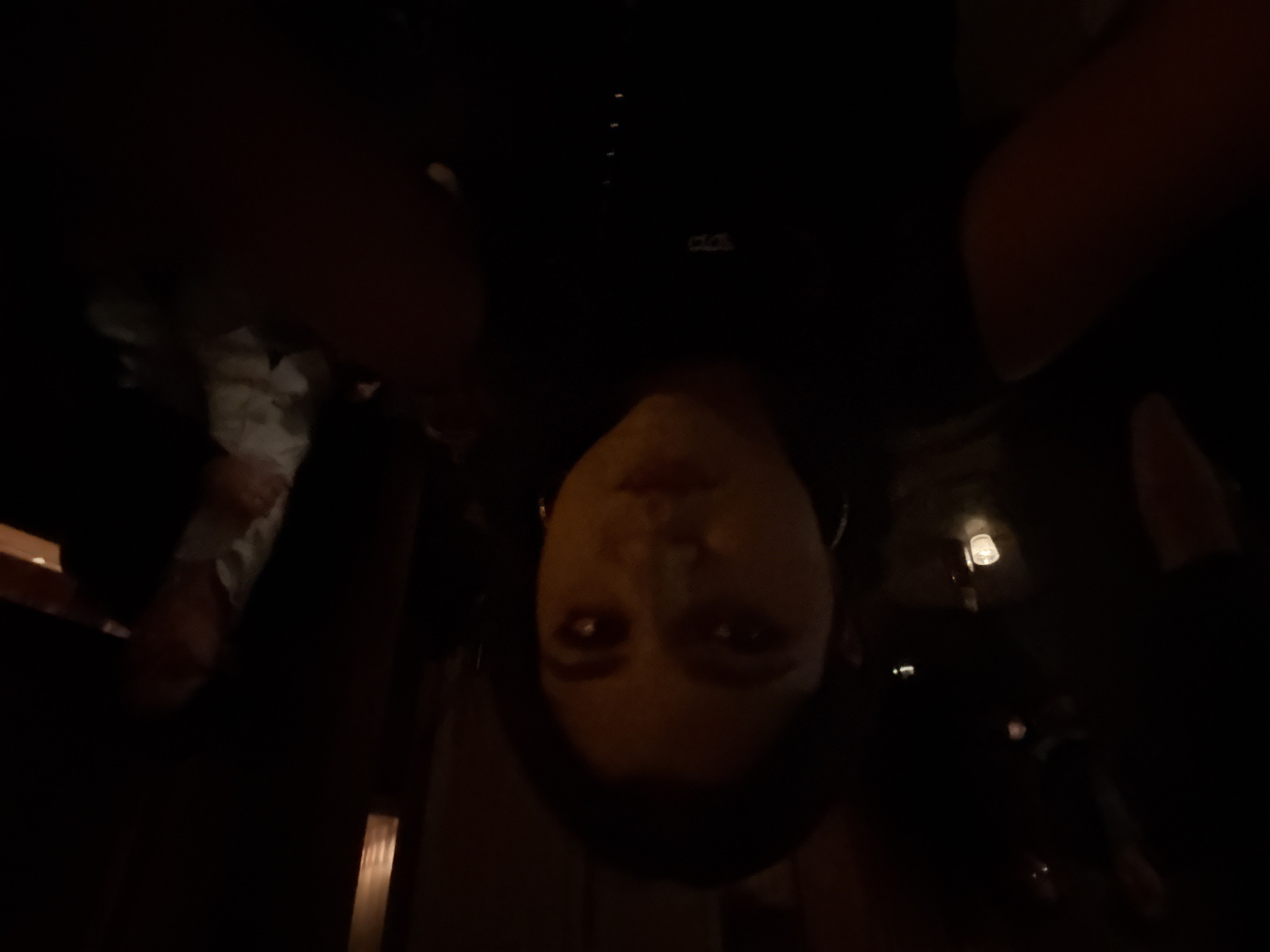 A person is seen upside down in a dimly lit indoor setting. The person has fair skin and dark hair. They are wearing earrings. The environment appears to be a restaurant or bar, with other people and objects visible in the dark background. The location might be in Mexico City, Mexico, based on the prompt.