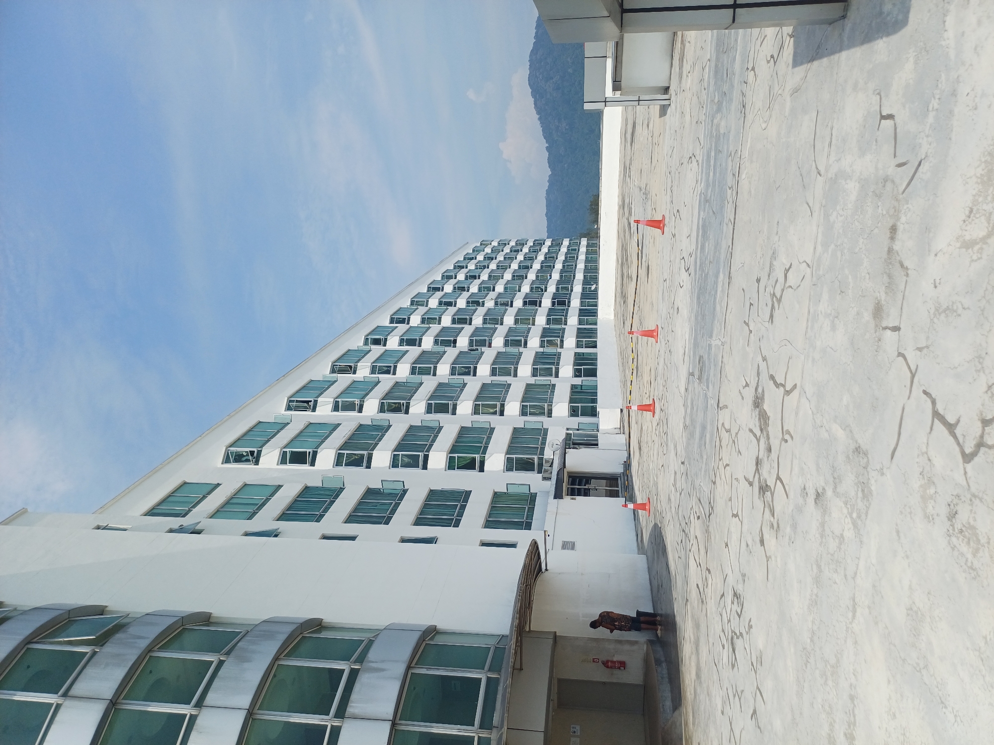 A long, white, multi-story building with many open windows stands under a bright blue sky with scattered clouds. In the foreground, a cracked concrete surface is marked with orange traffic cones. A person stands near the entrance of the building, looking at their phone. In the background, a green, forested mountain range is visible. The building appears to be an apartment complex or hotel, possibly located on the outskirts of George Town, Malaysia, given the tropical vegetation and architecture.