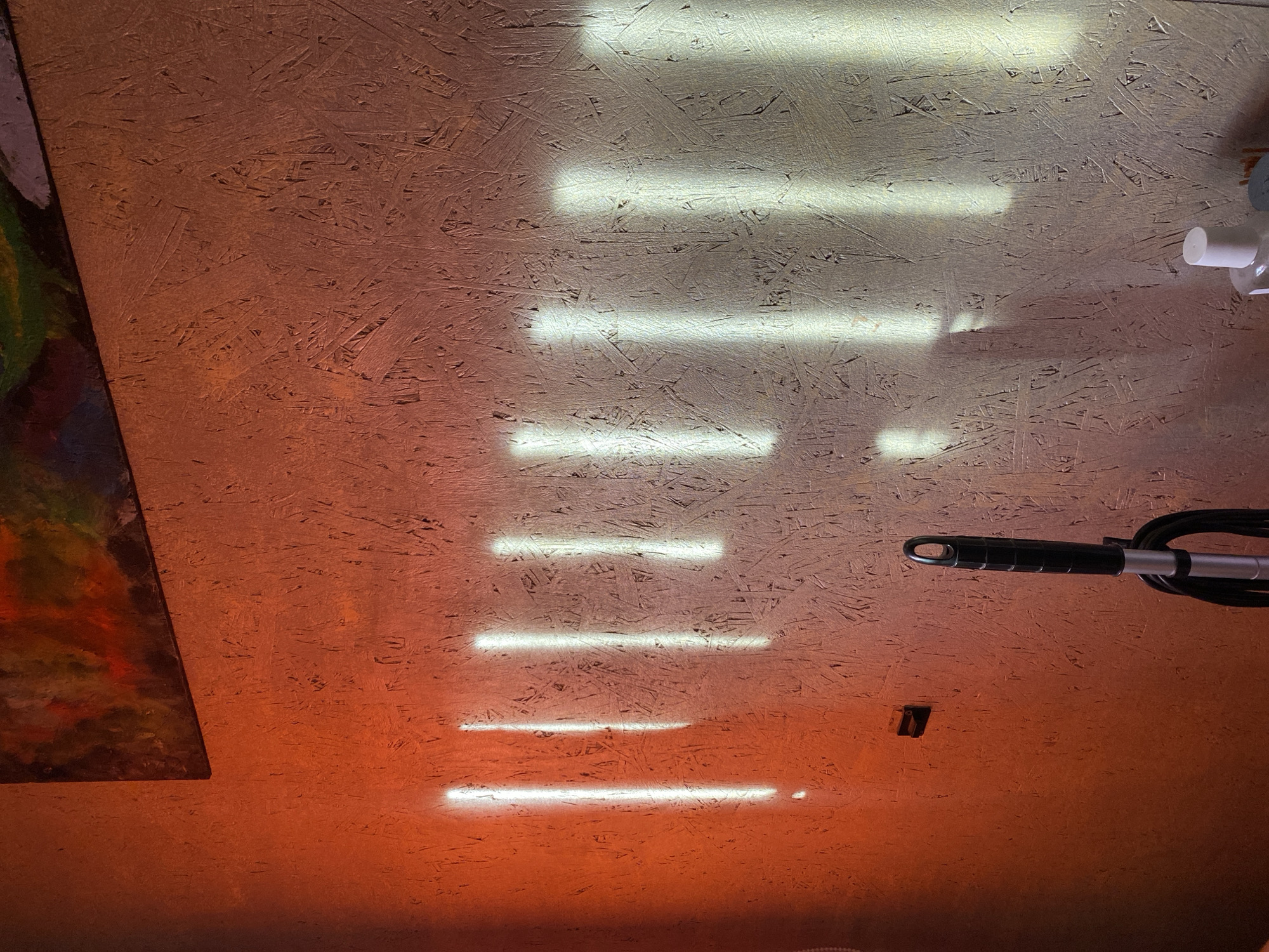 The image shows a section of a wall covered in what appears to be particle board, with uneven orange and gray coloring. The board features a pattern of long, thin wood chips. Six horizontal strips of light are shining on the wall, spaced irregularly. In the upper right corner, there is a white plastic dispenser. To the right, a black hose and silver rod are partially visible. On the left edge, there is a blurred section of what looks like a colorful painting with dark red, green, and brown hues. Near the bottom, there is a small, dark-colored object affixed to the wall. The location is Unknown, Unknown.